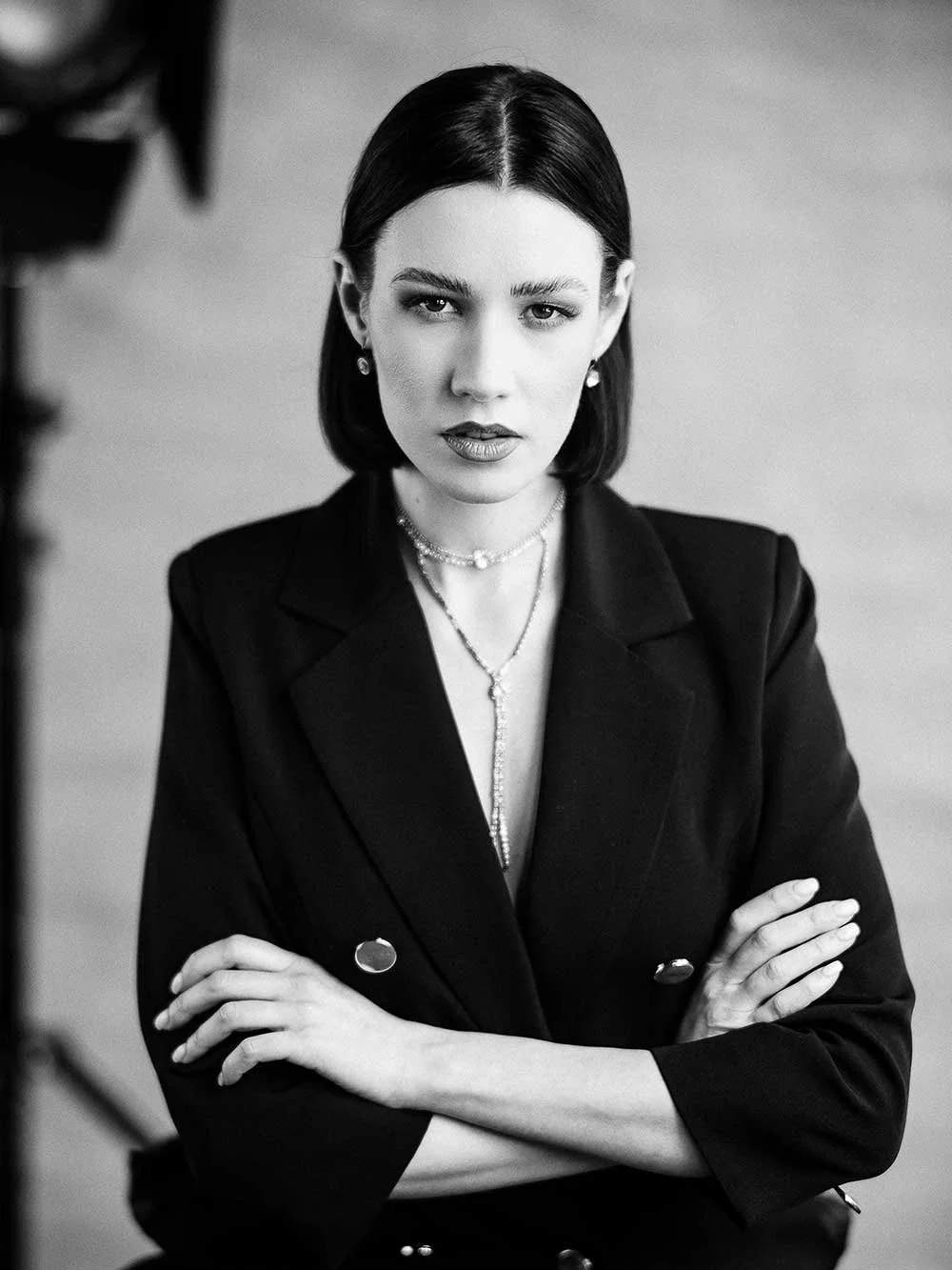 A black and white portrait of a woman with dark hair, wearing a dark blazer and layered necklaces, with arms crossed, looking directly at the camera.