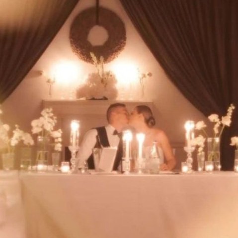 Una coppia seduta a un tavolo decorato con fiori e candele, ambientazione elegante con tende e decorazioni floreali.