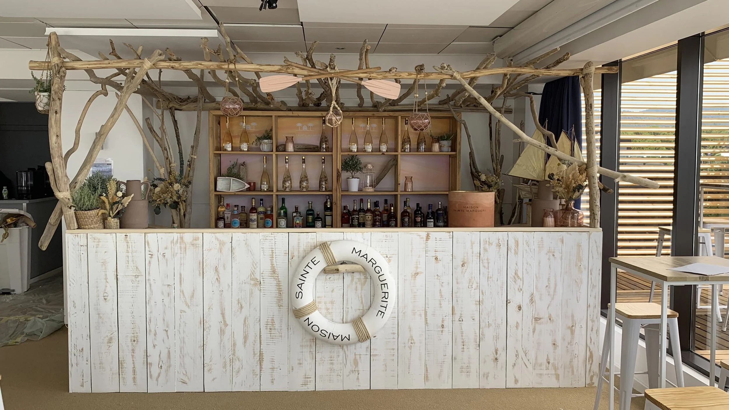 Un bar de style côtier avec un comptoir en bois blanc usé, décoré de plantes en pot et d'un cercle de vieilette avec la mention Sainte Marguerite. Derrière, des étagères en bois clair avec des bouteilles de différentes tailles et alcoolisées, ainsi que des décorations en bois et en verre. Des branches d'arbre décoratives forment une structure au-dessus du bar. À droite, des tables en bois avec des chaises en métal blanches, et une grande fenêtre avec des lames en bois lamellé laissant entrer la lumière naturelle.
