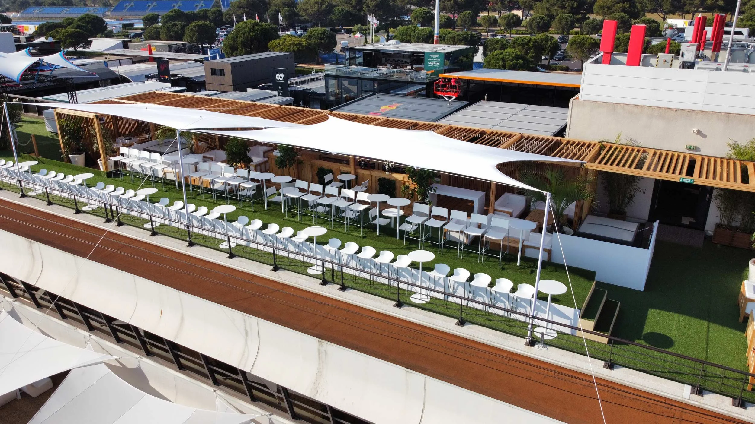 Rooftop lounge avec des tables hautes et des chaises blanches, un espace de détente avec des canapés, sous un auvent blanc, entouré de plantes. En arrière-plan, des bâtiments modernes et un parking.
