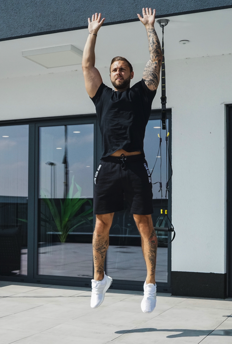 Mann beim Training an einem TRX-Gürtel, springt in die Luft und macht Klimmzüge im Freien.