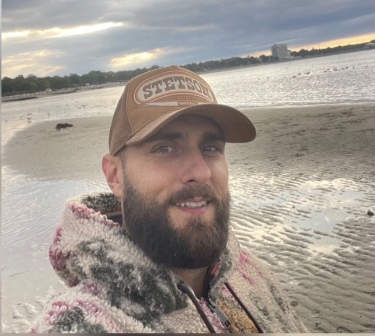 Ein Mann mit Vollbart trägt eine braune Cap mit der Aufschrift 'Cotton Stuff' und einen bunten, wolligen Pullover. Er steht an einem Strand, im Hintergrund Wasser, eine Fischreiher und ein bewölkter Himmel erkennbar.