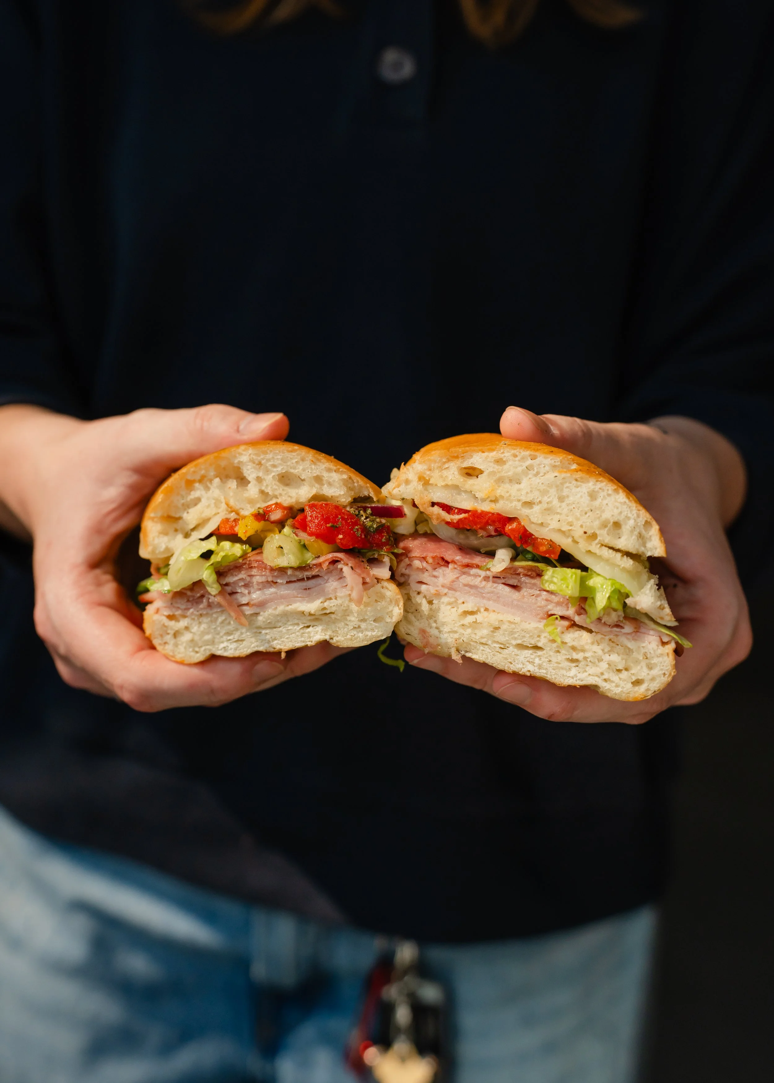 Person holding a sandwich with visible layers of garlic aioli, ham, prosciutto, salami, lettuce, roasted red peppers, banana peppers, red onion, toasted on a fresh hoagie