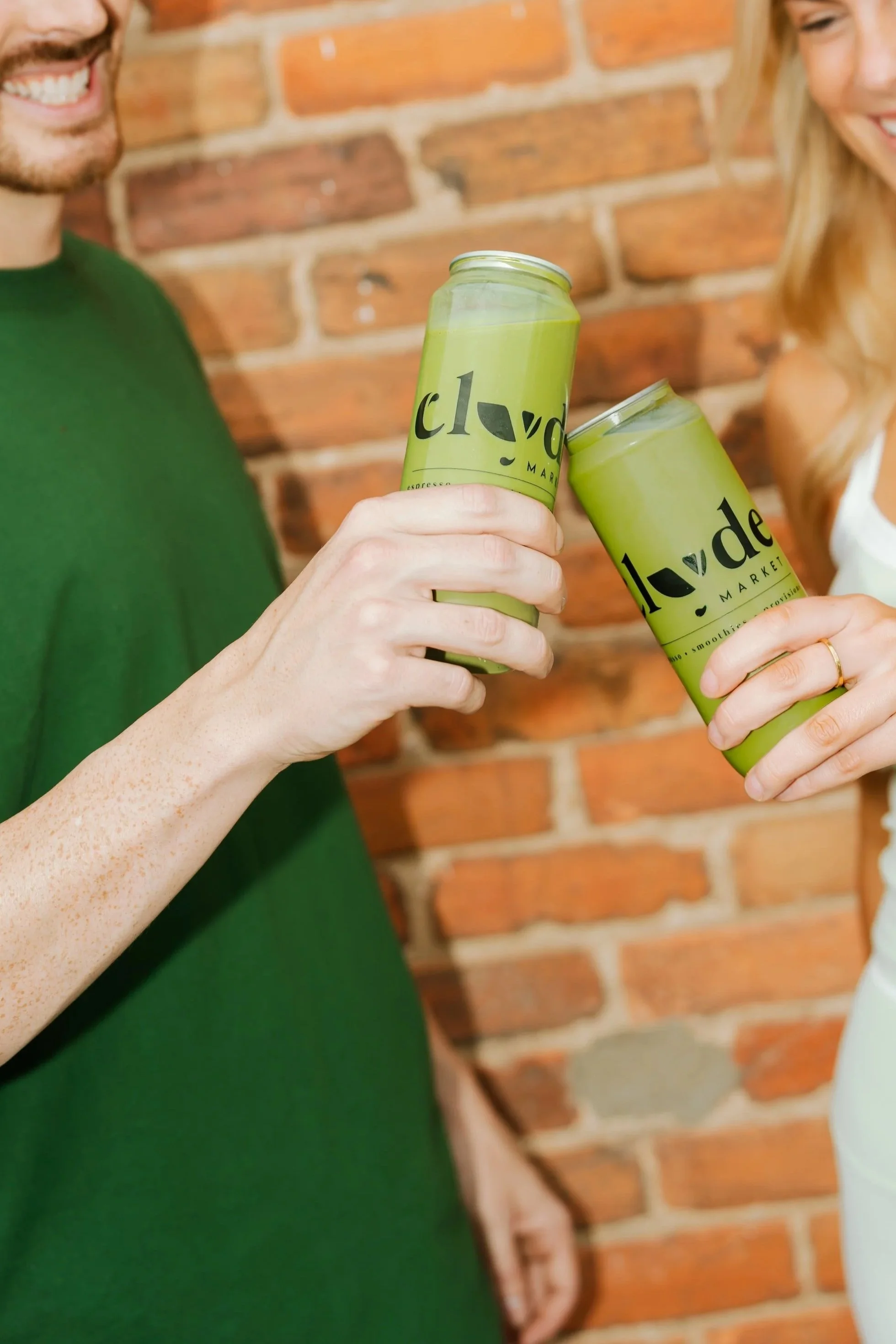 Two canned matcha drinks being clinked together in a celebratory “cheers.” The cans are chilled and slightly frosty, suggesting a refreshing beverage, with a bright green matcha color visible on the packaging and a fun, casual social moment.