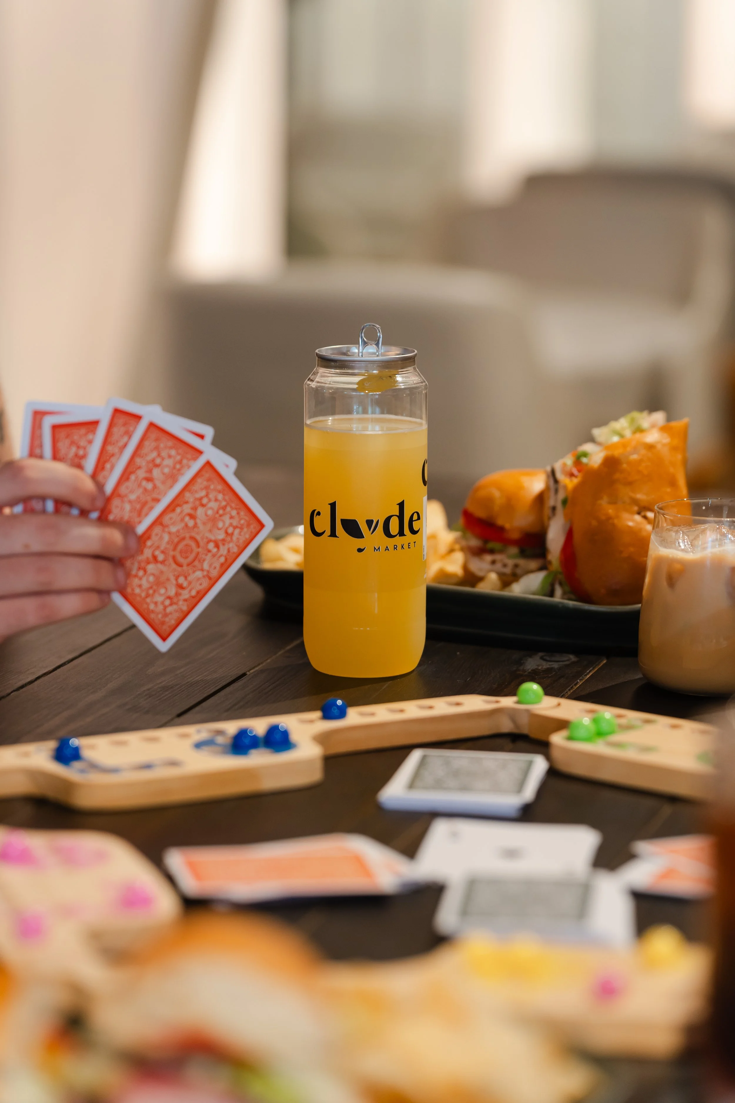 Close-up of a table with playing cards, a glass of juice, and a plate of sandwiches. In the background, a person is holding a set of red-backed playing cards. The focus is on the playing cards, with the rest of the scene slightly blurred.