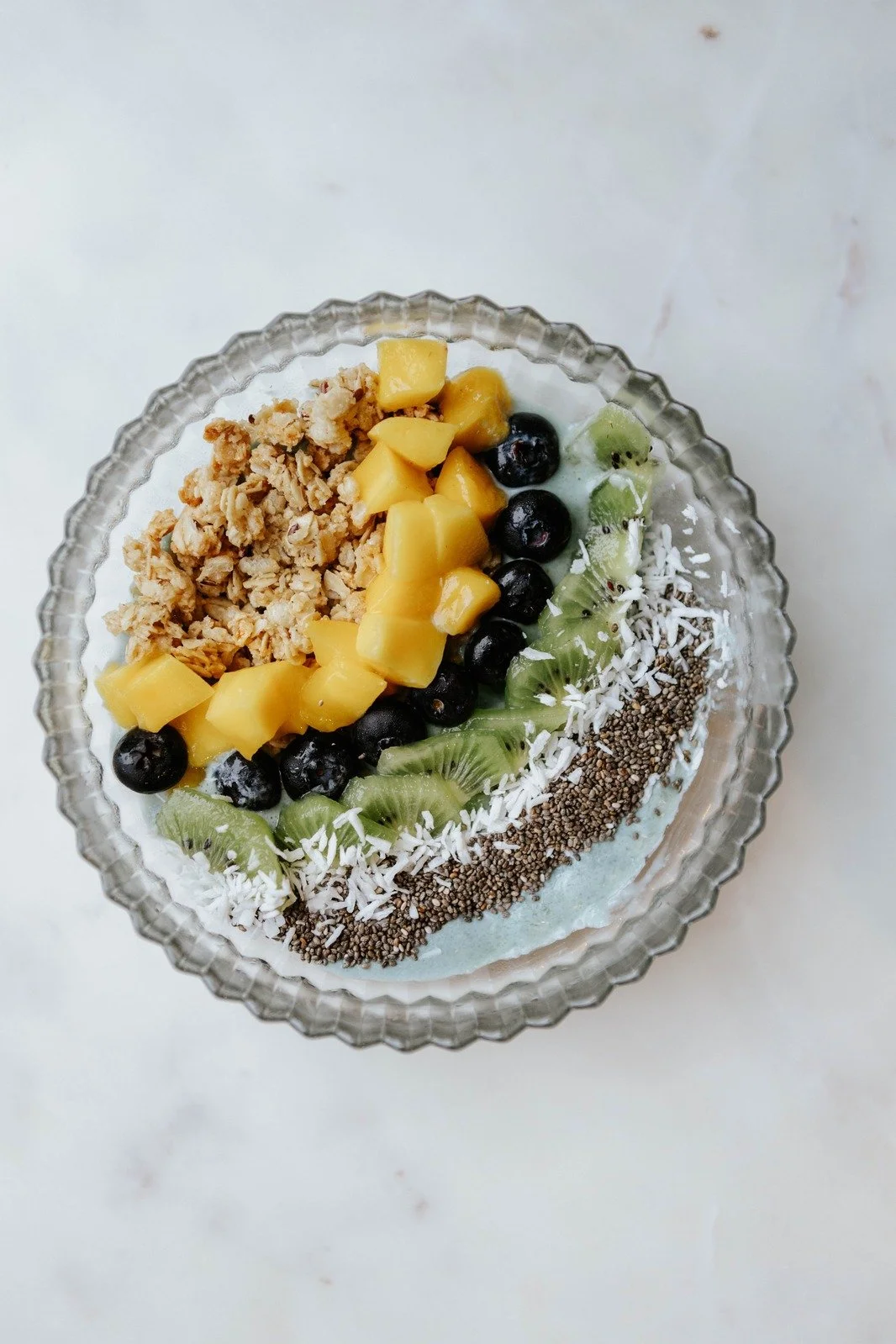 A coconut cream–blended bowl with a smooth, creamy white base, topped with coconut flakes, sliced kiwi, blueberries, mango chunks, and crunchy gluten-free granola. The bowl is brightly colored and tropical.
