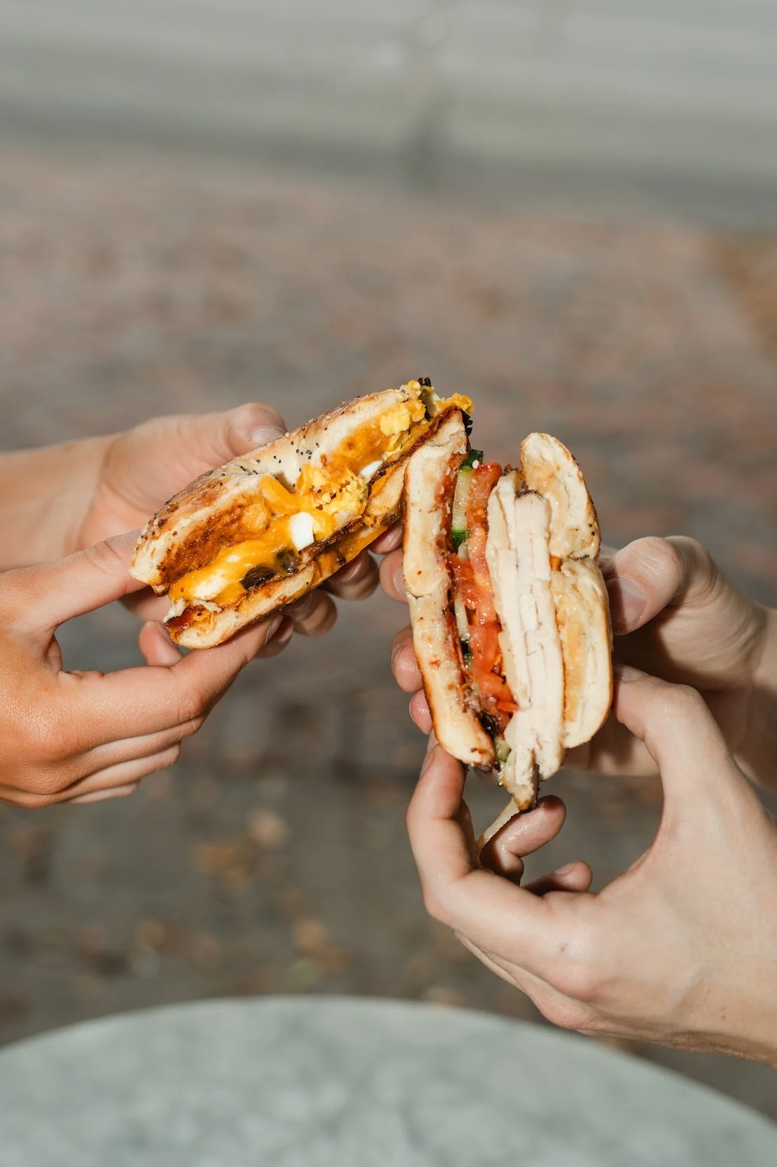Two hands holding halves of a breakfast sandwich with melted cheese, egg, chicken, tomato, and green onion, outdoors with a blurred background.