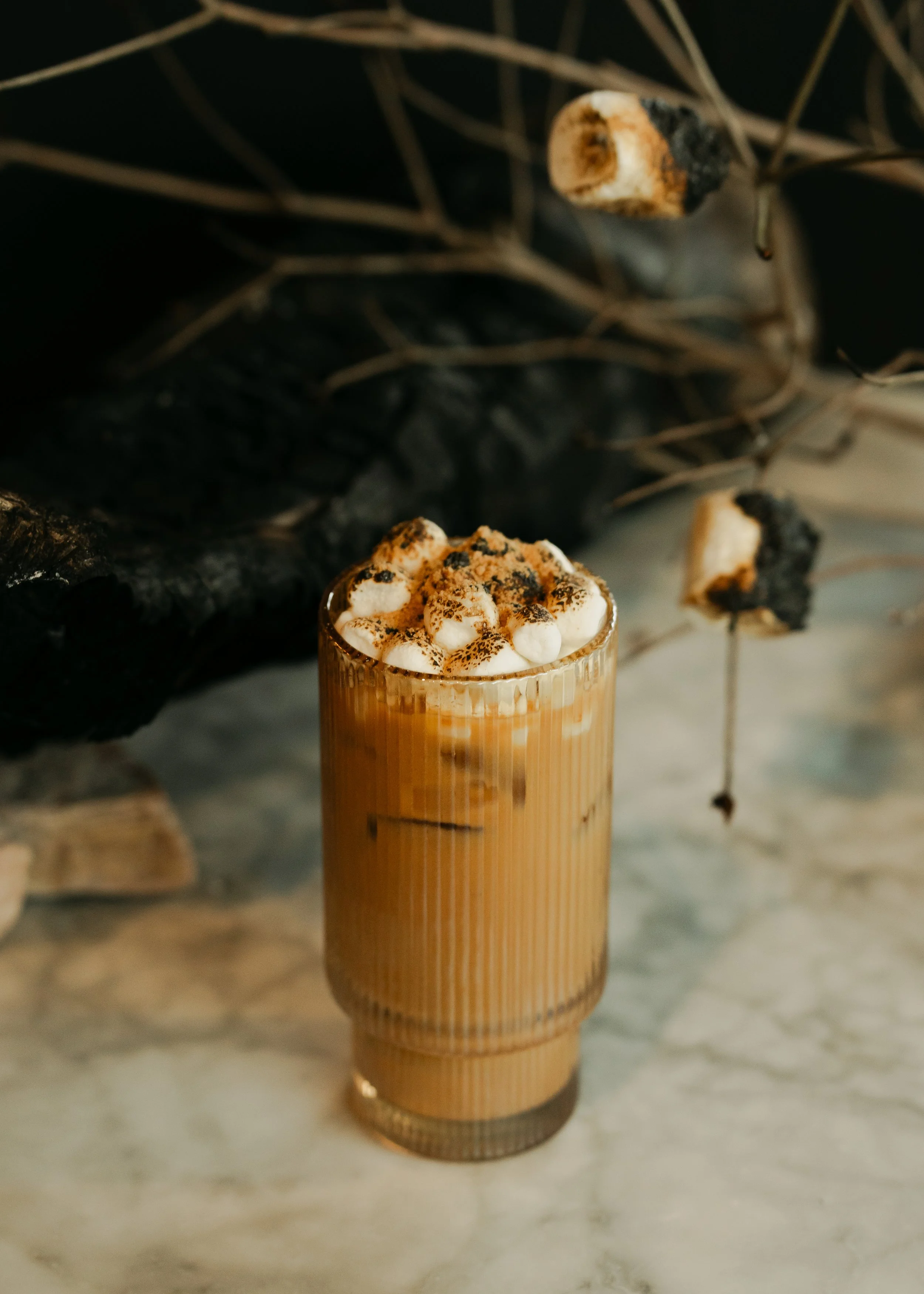A tall glass filled with sweet potato syrup, topped with marshmallows, and graham cracker with dried twigs or branches in the background.