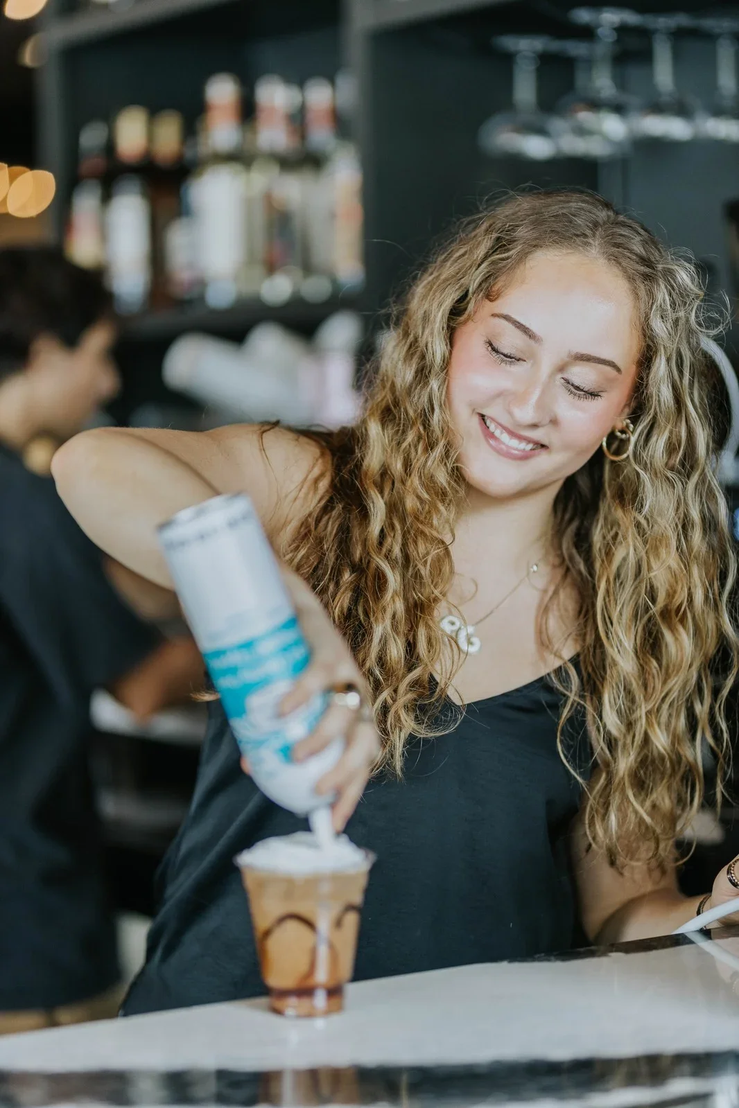 A woman with curly blonde hair smiling as she pours whipped cream on a mocha latte in a coffee shop.