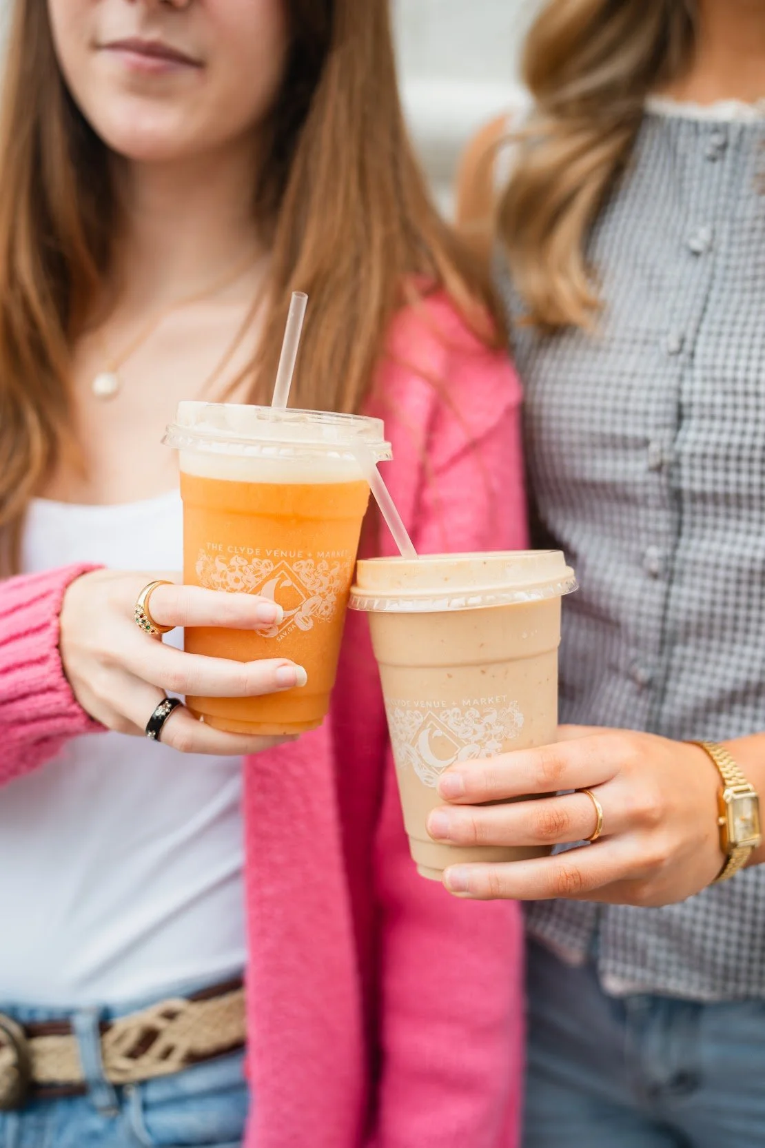 Two women holding iced beverages in disposable cups, one with an tropical smoothie and the other with a peanut butter chocolate smoothie drink, both with straws.