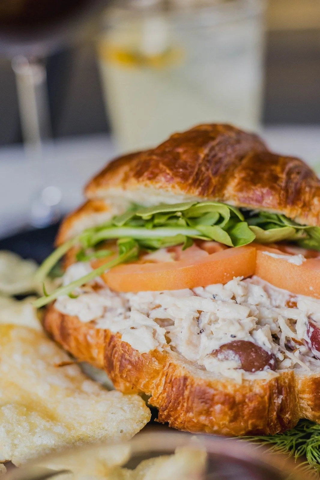 A chicken salad sandwich made with housemade chicken salad mixed with mayo, ranch, grapes, and celery, served on a fresh croissant or sourdough. The sandwich is layered with romaine lettuce and sliced tomatoes,