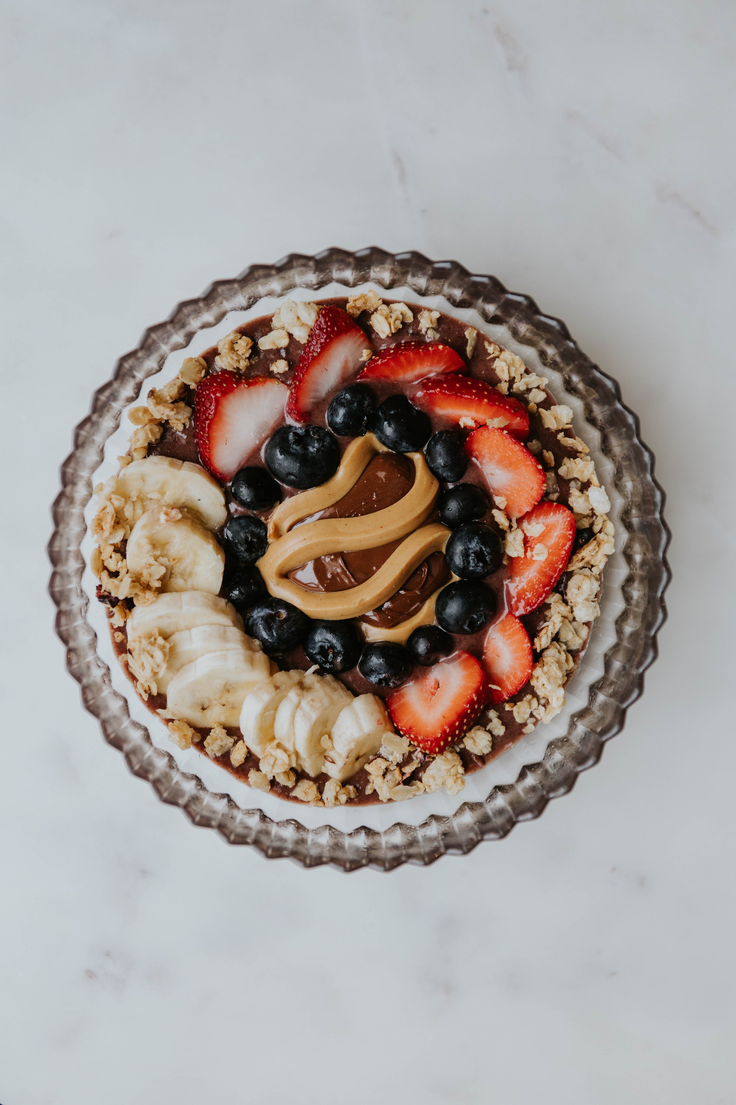 A rich açaí bowl blended with chocolate plant-based protein and topped with creamy peanut butter, hazelnut cocoa butter, fresh blueberries, sliced strawberries, banana, and gluten-free crunchy granola.