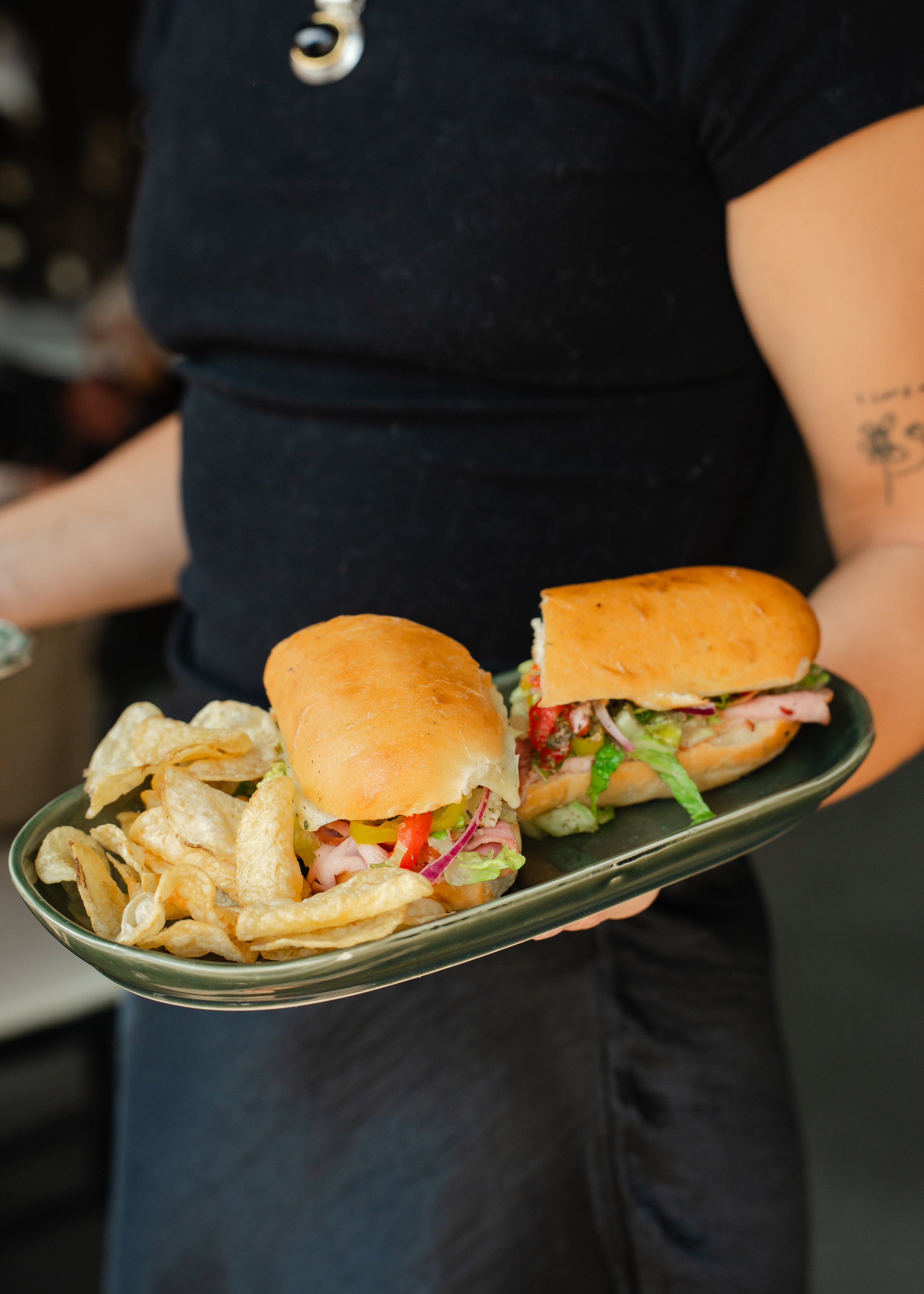 Person holding a sandwich filled with garlic aioli, ham, prosciutto, salami, lettuce, roasted red peppers, banana peppers, red onion, toasted on a fresh hoagie and potato chips. 
