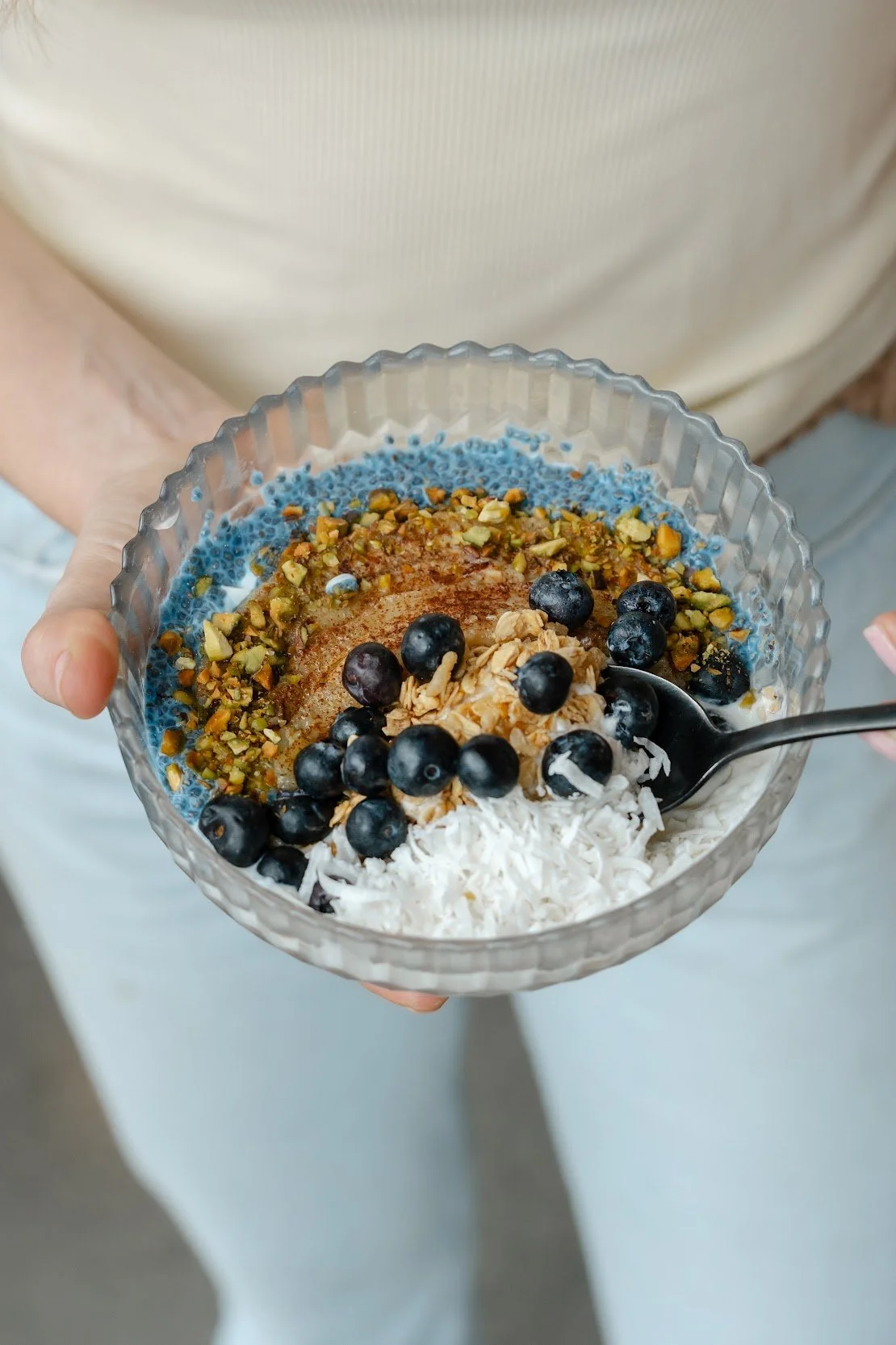 A vanilla yogurt bowl topped with vibrant blue spirulina chia seed pudding, roasted cinnamon-salted banana slices, crunchy granola, fresh blueberries, chopped pistachios, and coconut flakes.