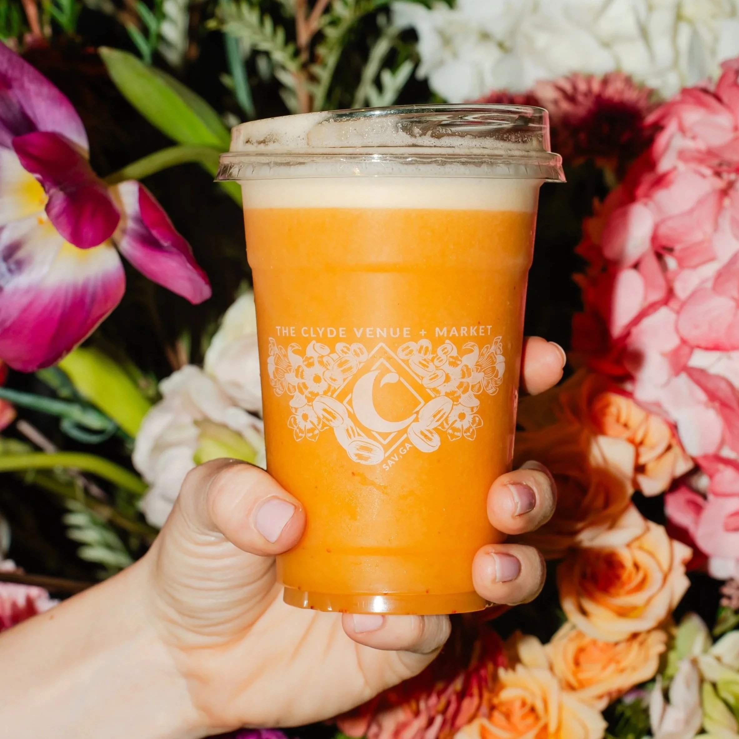 A hand holding an orange-colored beverage in a clear plastic cup filled with mango, pineapple, strawberry, banana, lemon, orange juice