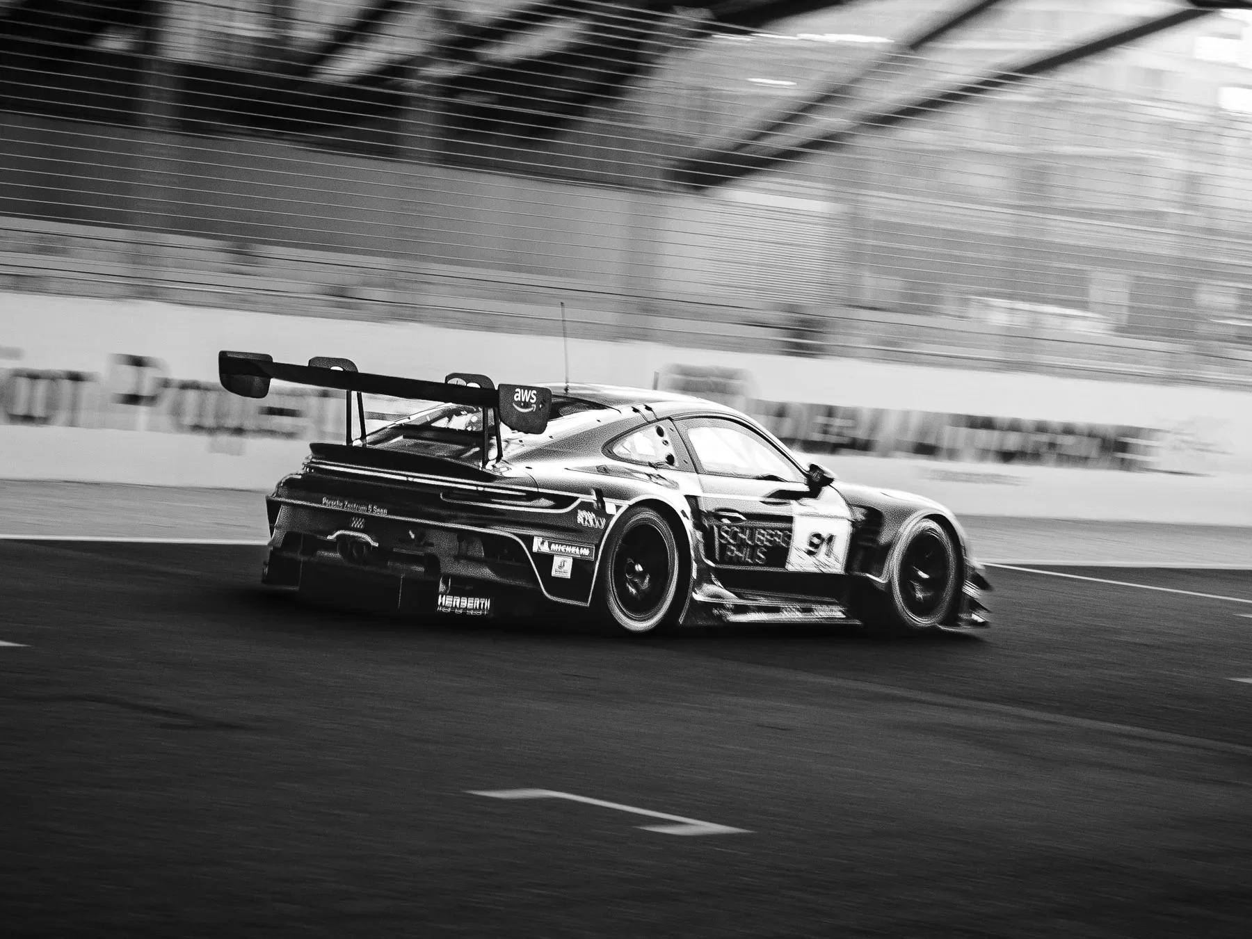 Black and white photo of a race car speeding on the track with a large rear wing and sponsor logos, indoors at a racing venue.