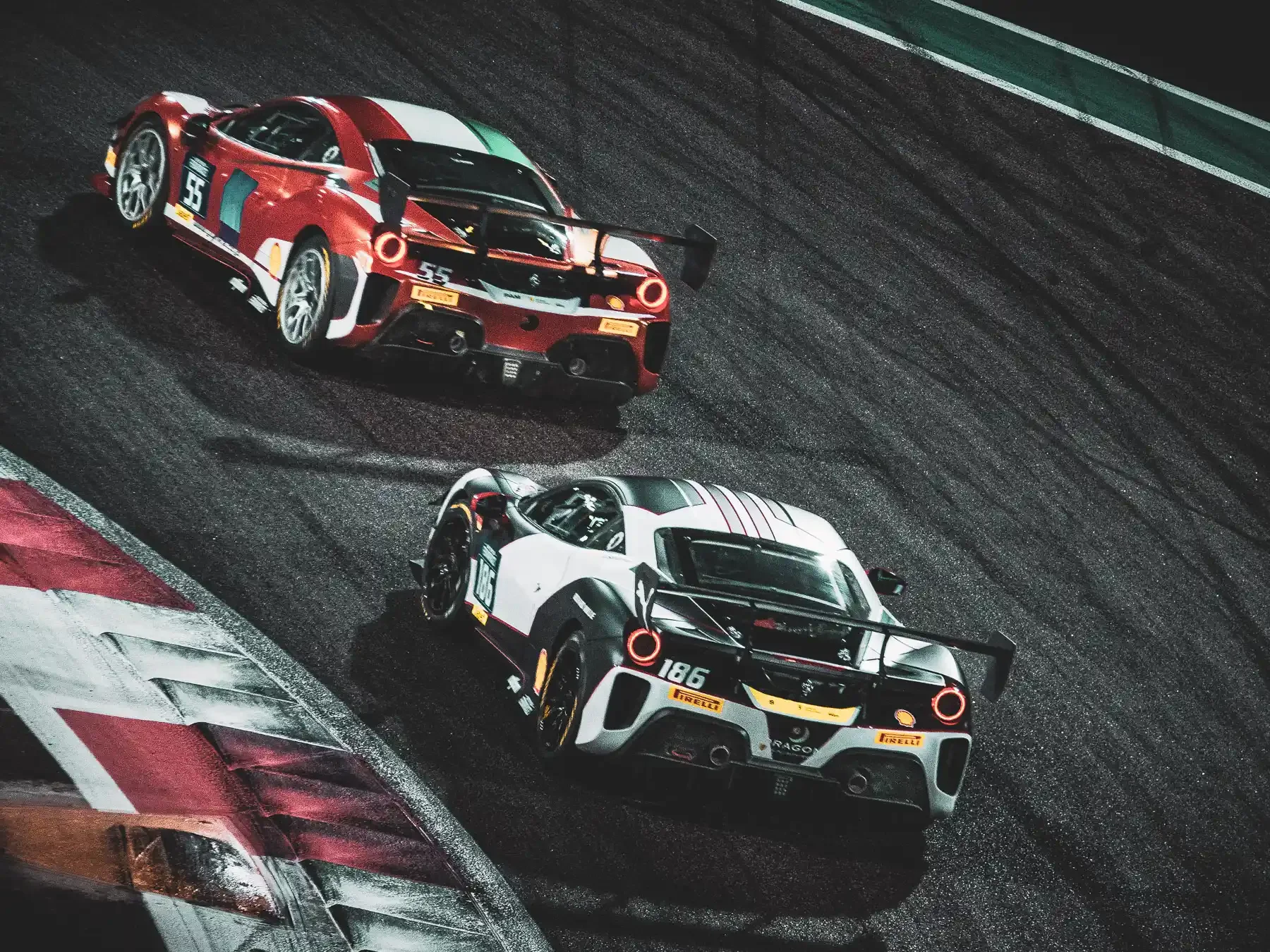 Two race cars on a track at night, one red with white accents and the other black and white, racing closely.