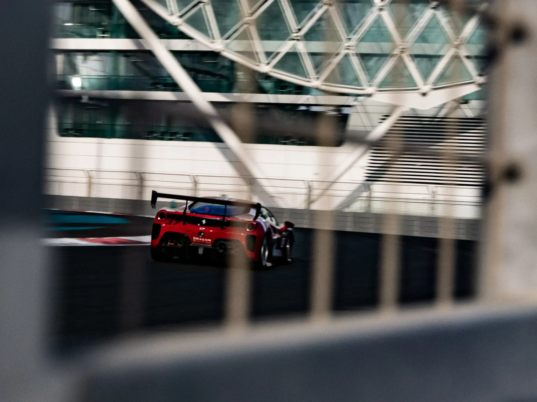 A red race car on a track, seen through a metal fence and in front of a modern glass building.