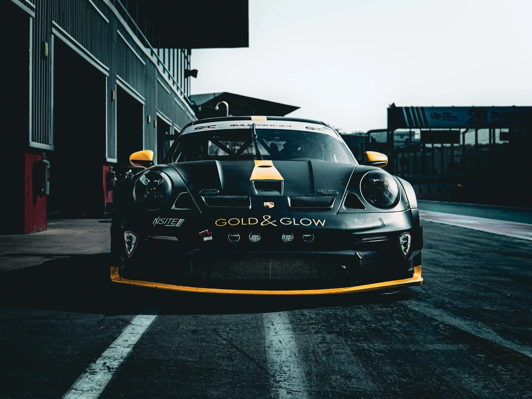Black race car with yellow accents parked in a pit lane at a race track, with pit garage and race track in the background.