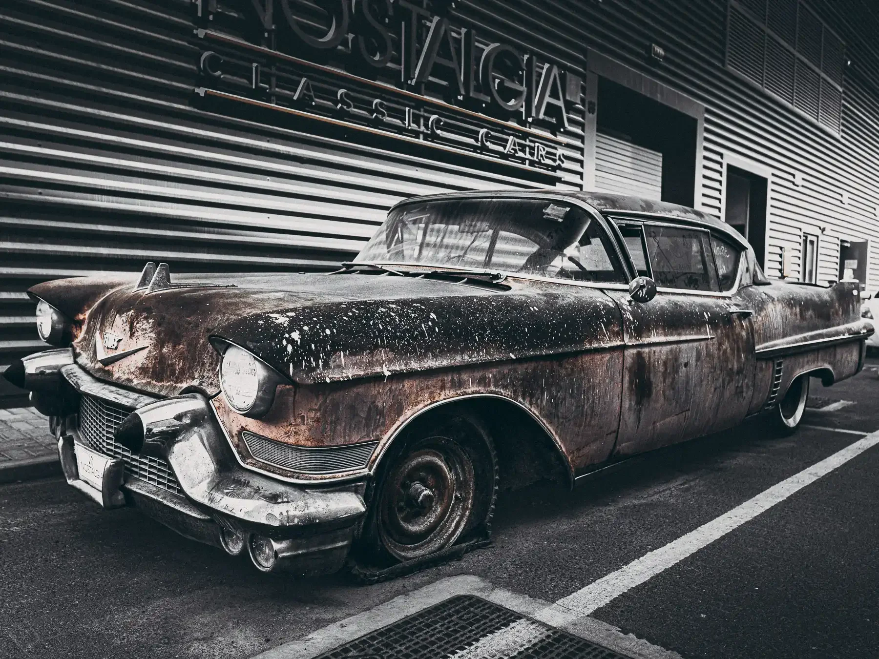 Old rusty vintage car parked outside a modern building with signage reading 'STAGIA' and 'CHASSIS'.