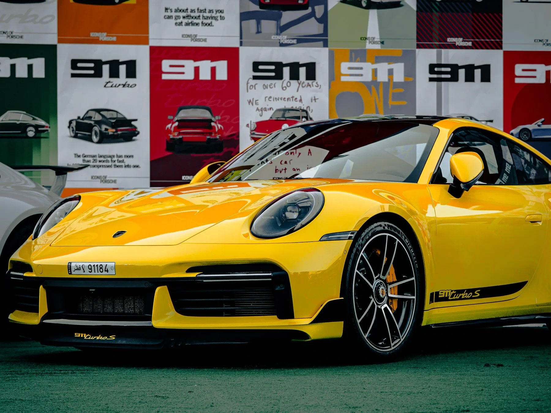 A yellow Porsche 911 Turbo S sports car parked at an auto show with colorful posters and cars in the background.