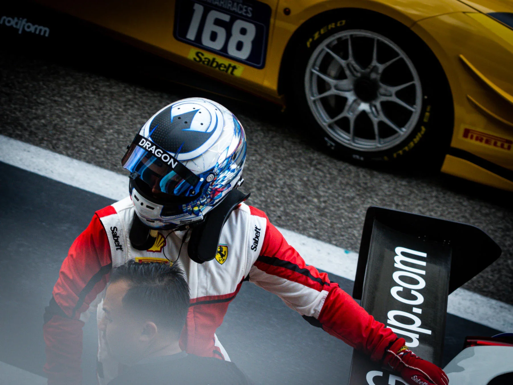 Close-up of a racing driver in a white and red suit and a blue helmet, leaning into a pit stop beside a black tire and a yellow race car with a number 168 and websites on the side.