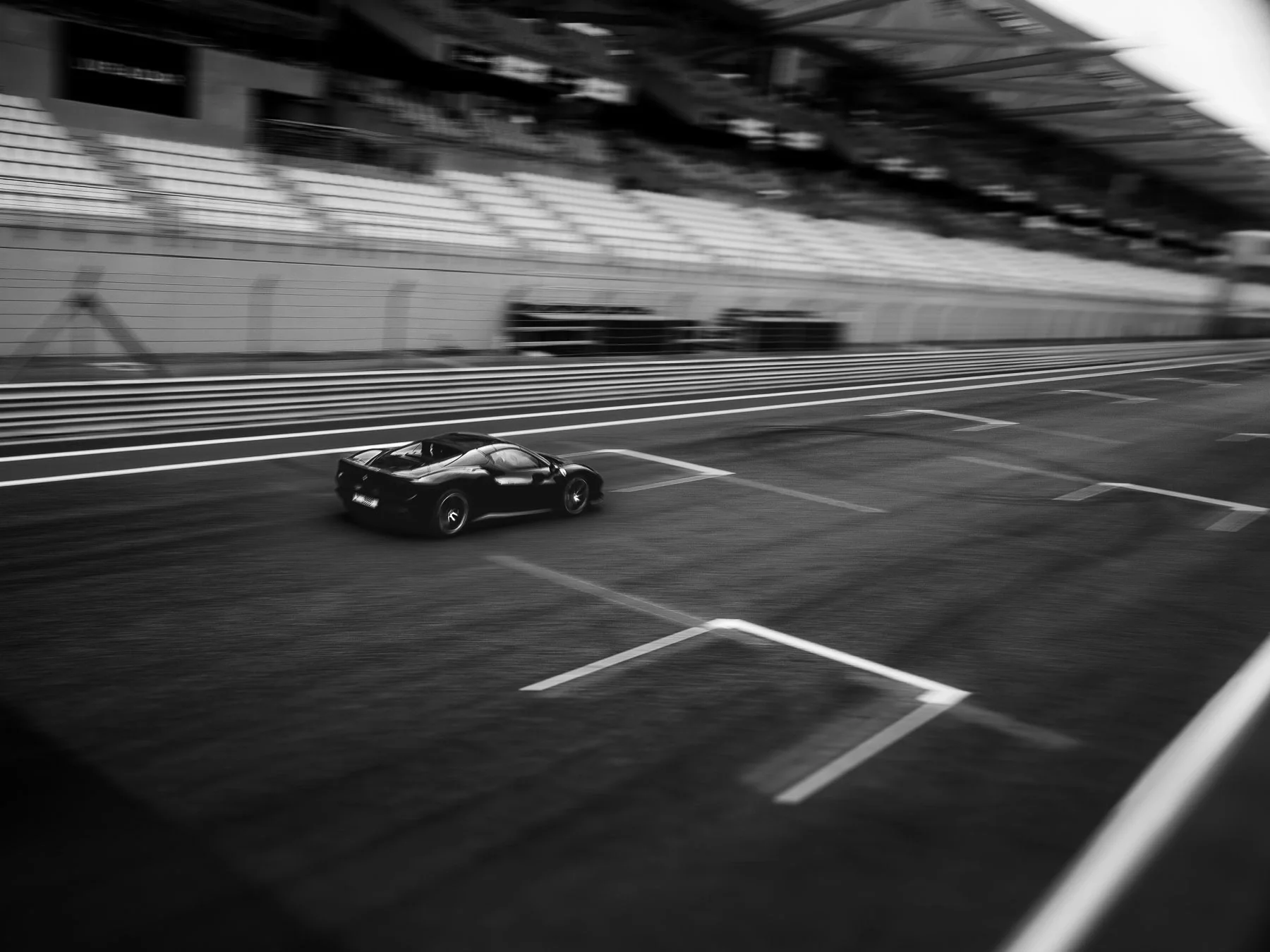 A black sports car moving quickly in an empty race track with grandstands in the background, captured in black and white with motion blur.