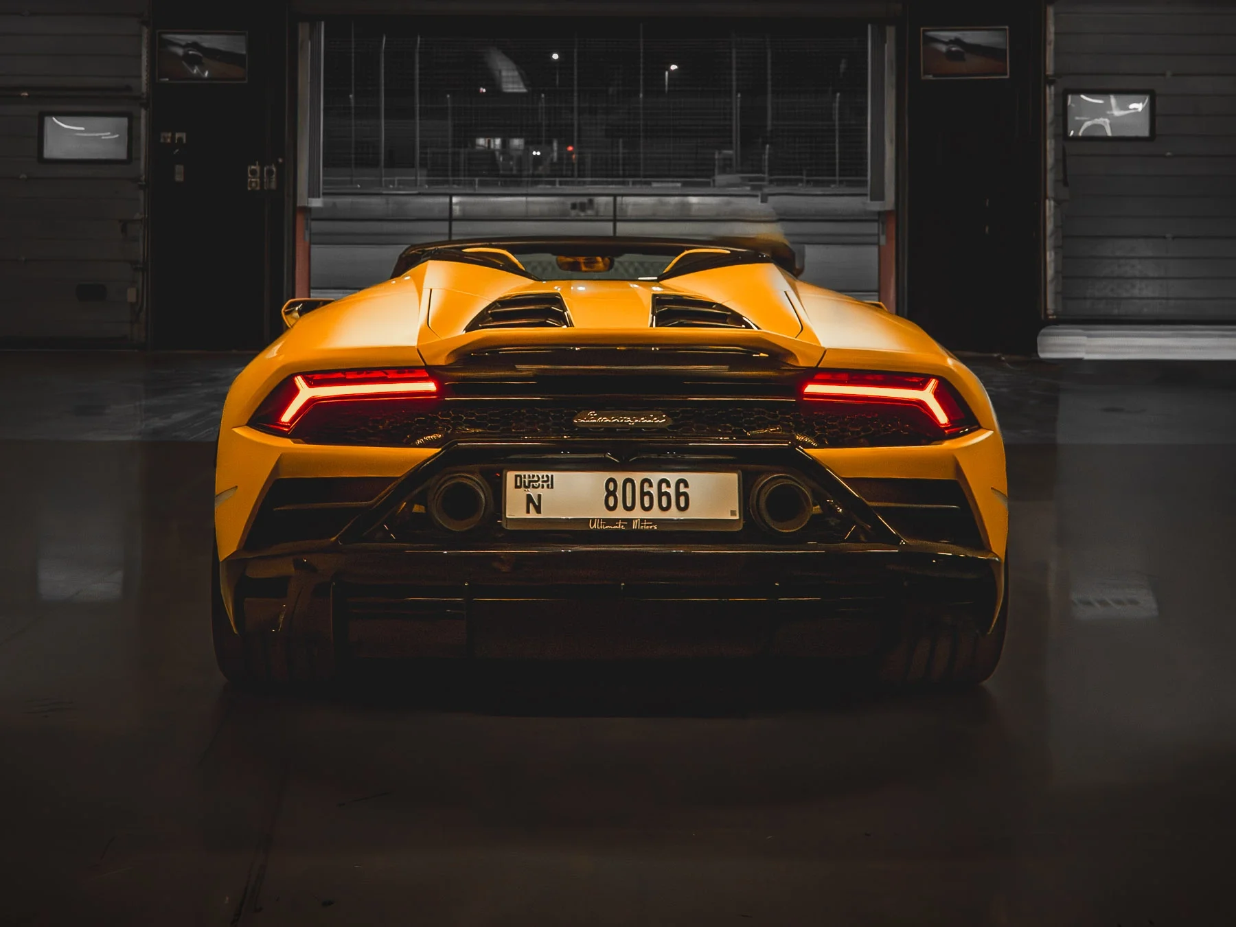 Yellow Lamborghini sports car parked in a garage with illuminated taillights, rear view showing dual exhausts and a Dubai license plate.
