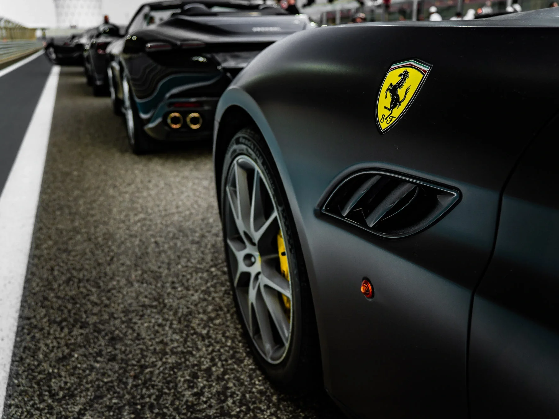 A close-up of a black Ferrari with a yellow prancing horse emblem, parked on a racetrack with other luxury cars in the background.