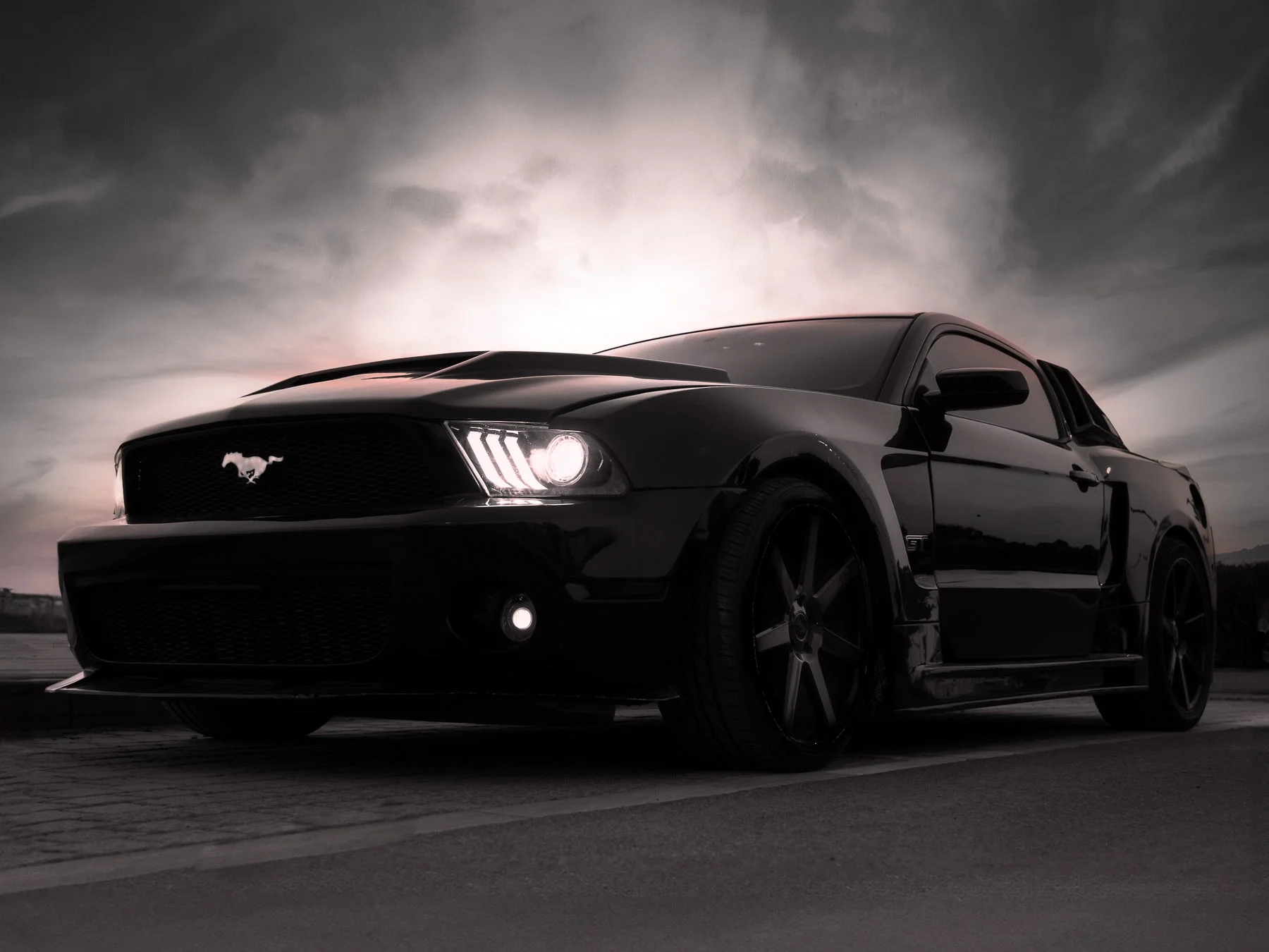 A black Ford Mustang sports car parked on a road during sunset or sunrise with cloudy sky.