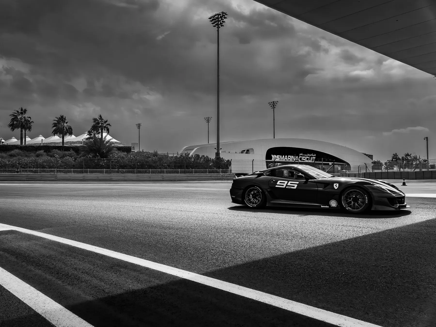 A black sports car with racing decals on a race track with a racing circuit building in the background, palm trees, and dramatic cloudy sky.