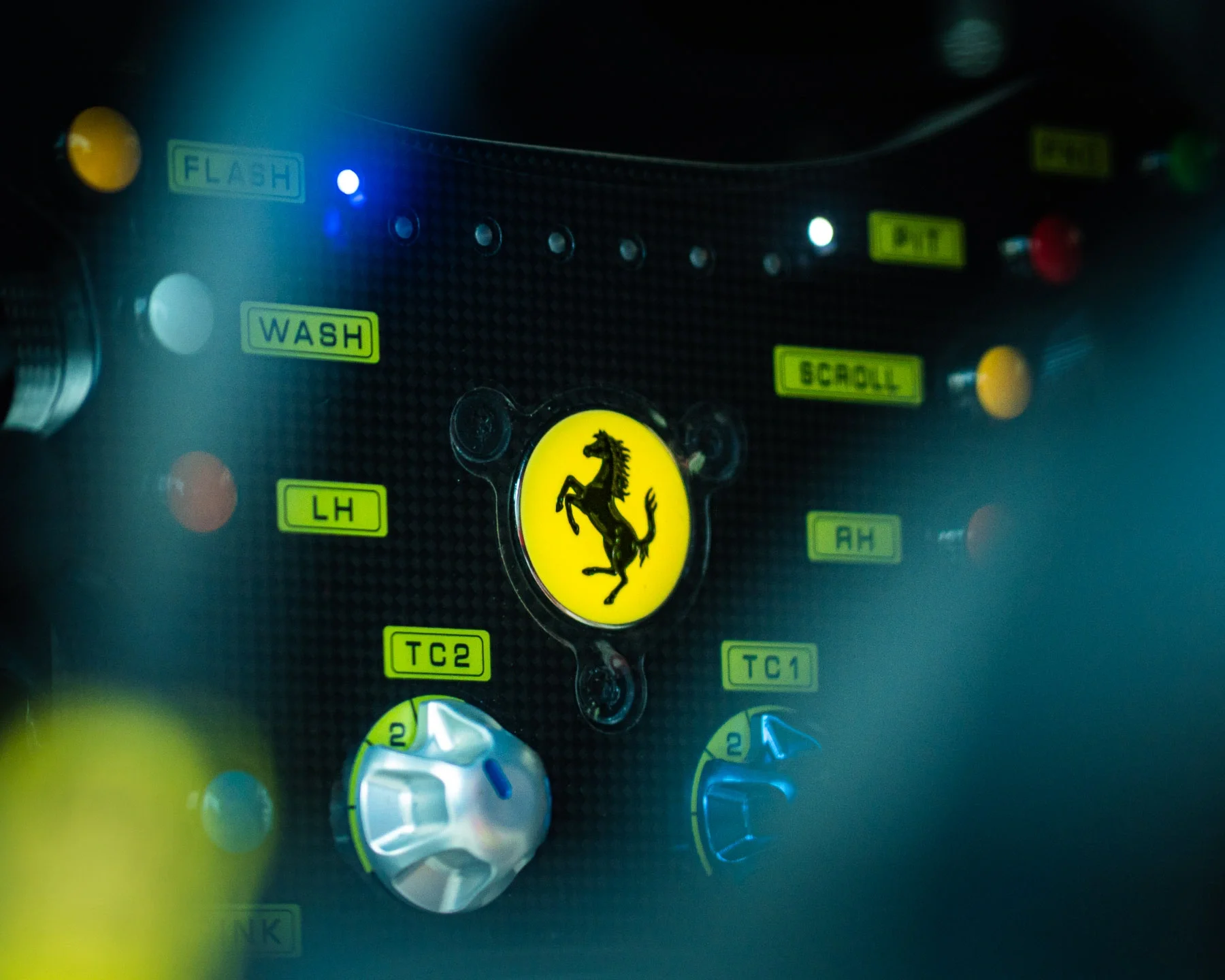 Close-up of a Ferrari F1 steering wheel with yellow prancing horse logo in the center, surrounded by various buttons, switches, and labels including 'WASH', 'SCROLL', and 'T1'.
