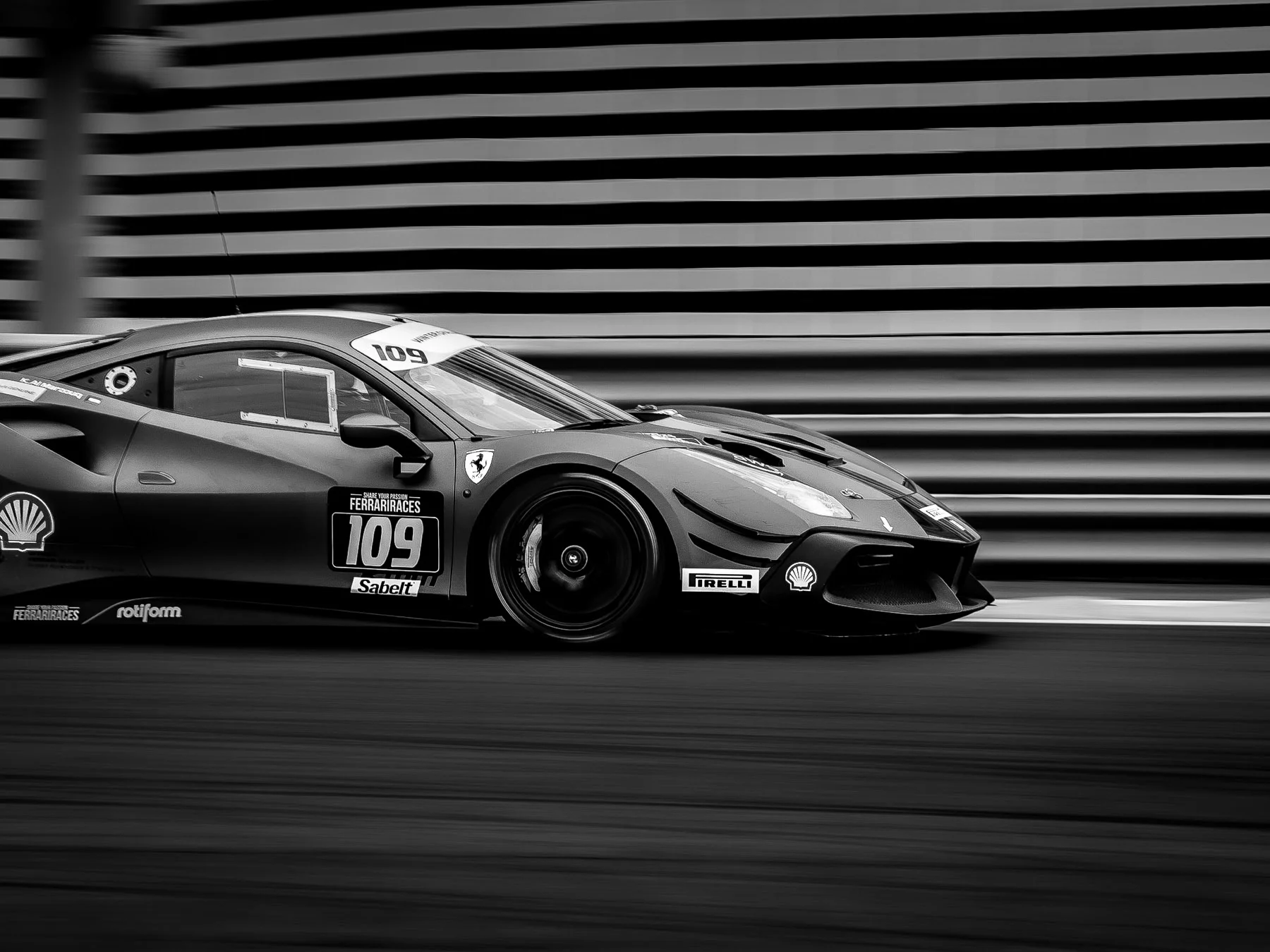 A black and white photograph of a racing Ferrari car on a track, number 109, with sponsor logos, in motion with blurred background.