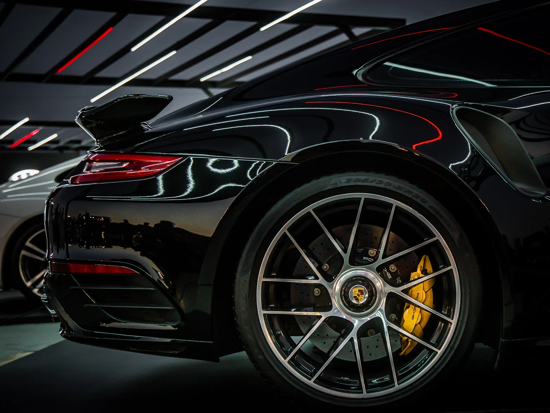 Close-up of a black Porsche sports car's rear wheel, brake caliper, and rear spoiler, parked inside a showroom with reflective lighting.