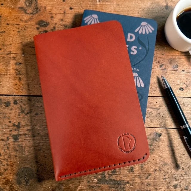 Cognac full grain leather Field Notes sheath by J.A.B. Leatherworks on rustic wood with a Field Notes notebook,