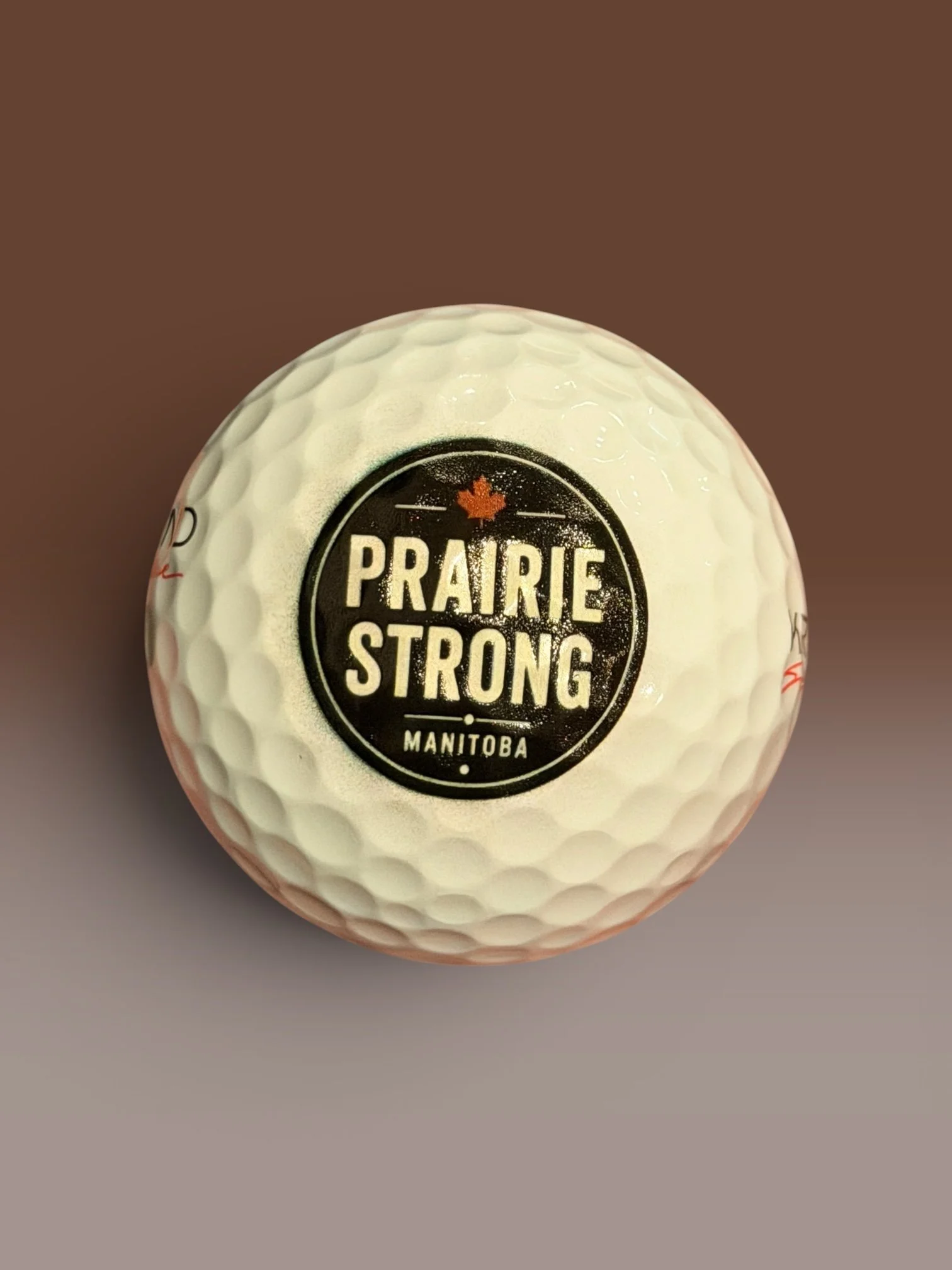 White golf ball printed with a bold circular badge design reading "Prairie Strong — Manitoba" with a red maple leaf