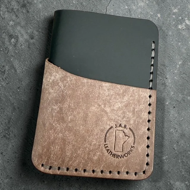 Two-tone full grain leather vertical card holder by J.A.B. Leatherworks, featuring smooth black upper and distressed grey lower panel with hand-stitching, on a grey stone surface.