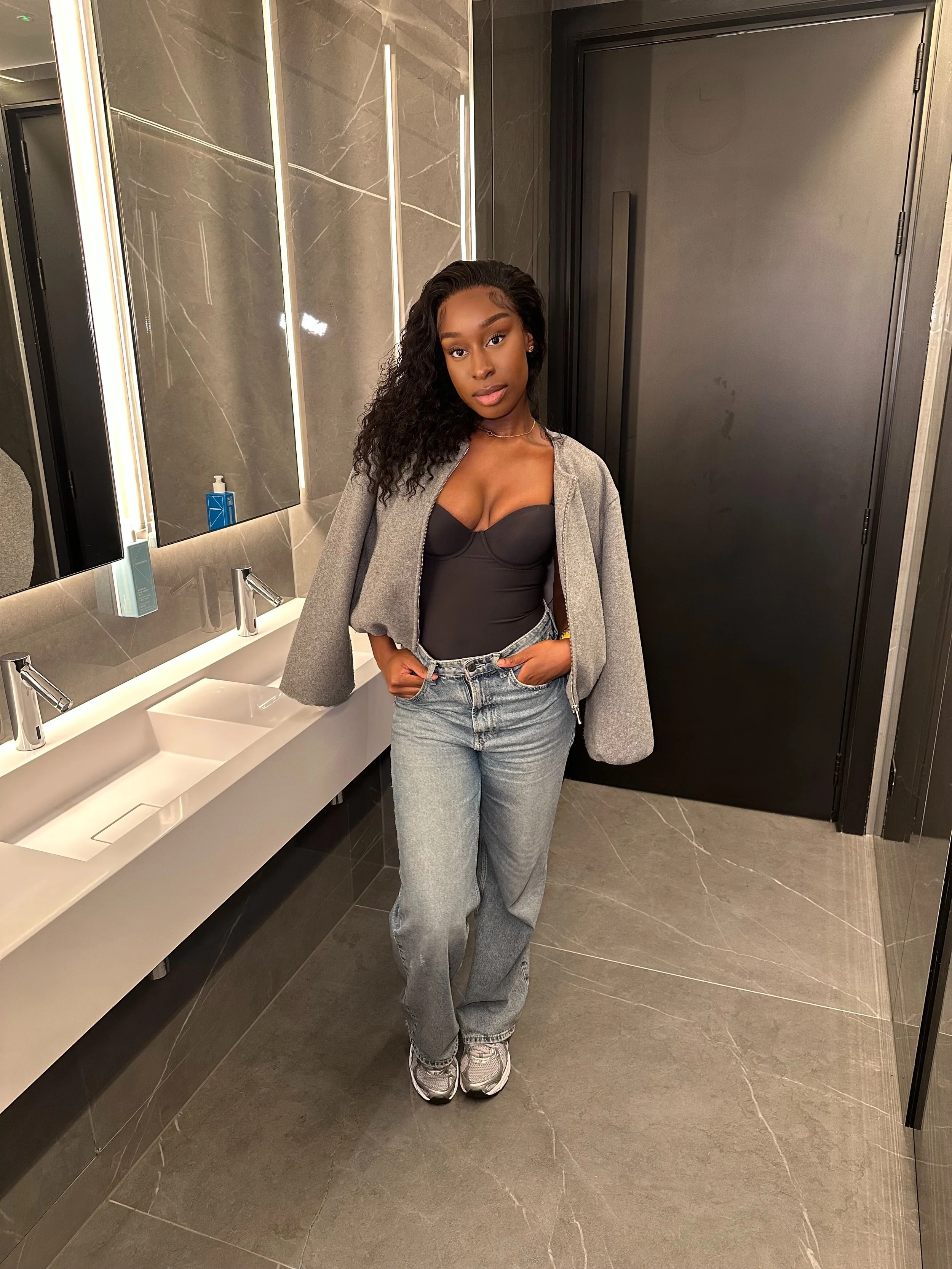 Young woman standing in a modern bathroom with gray marble walls and a double sink, wearing a black top, gray jacket draped over her shoulders, light denim jeans, and sneakers.