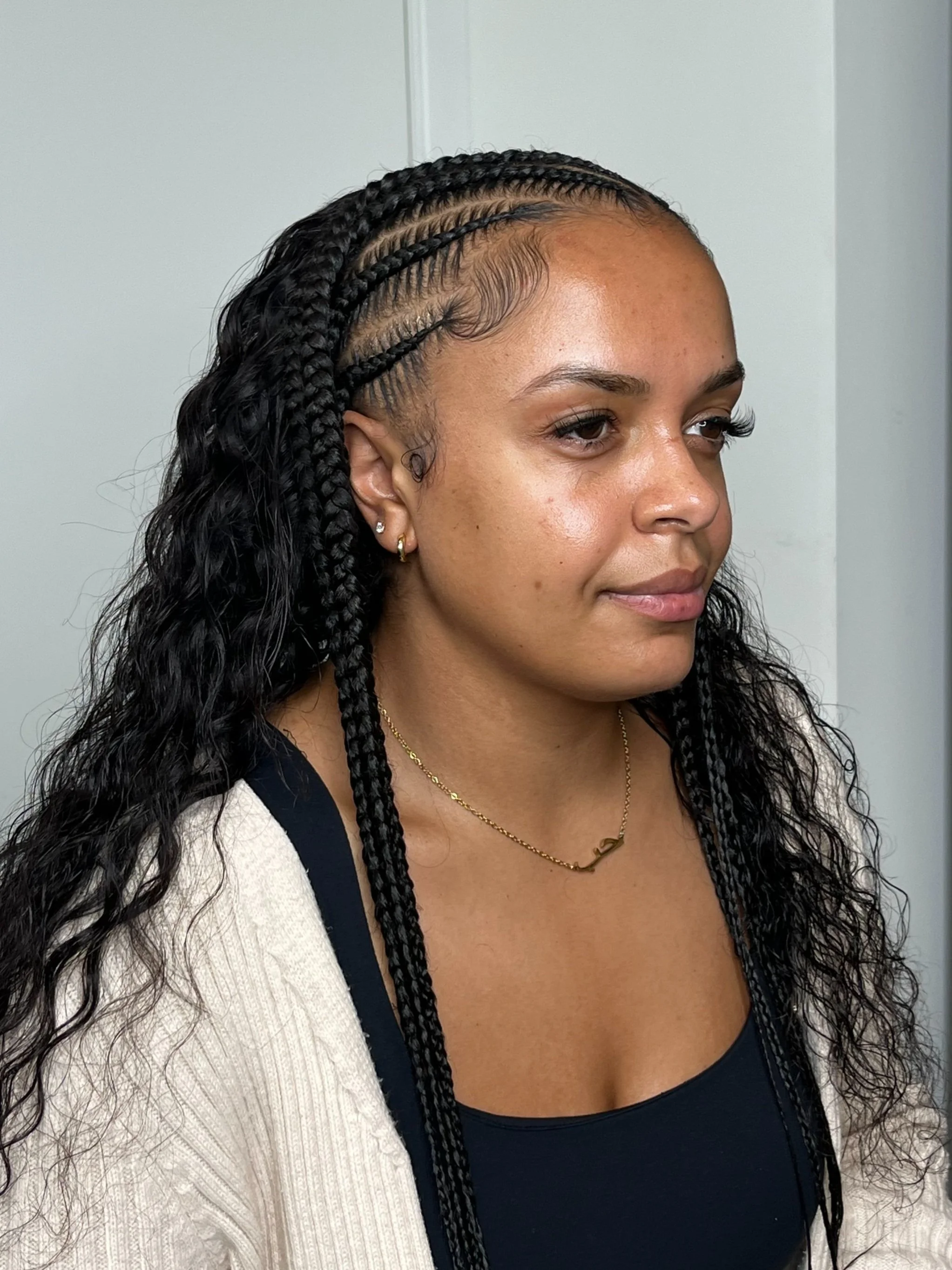 A woman with medium brown skin and long black curly hair styled in box braids. She is wearing a black top, a beige cardigan, and gold jewelry, including earrings and a necklace. She appears to be indoors, near a white wall.
