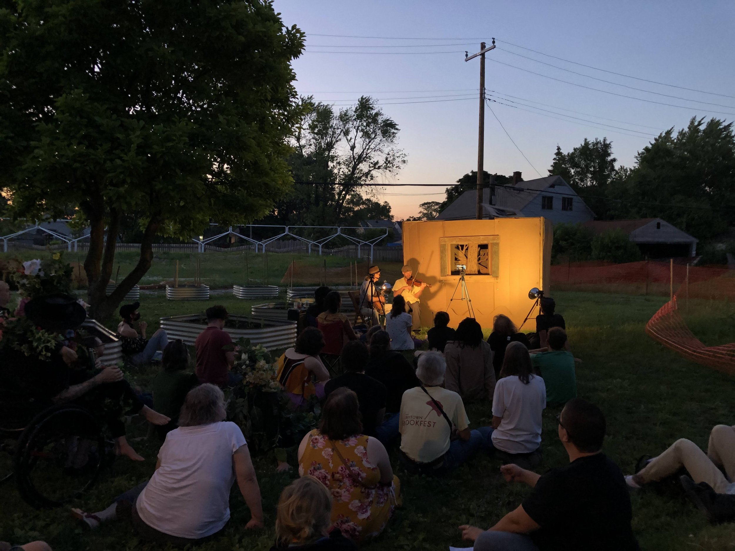 An evening outdoor performance with a cardboard stage and backdrop, as part of the annual Detroit Puppet Slam, with an engaged seated audience. Live music by two musicians accompanies the performance.
