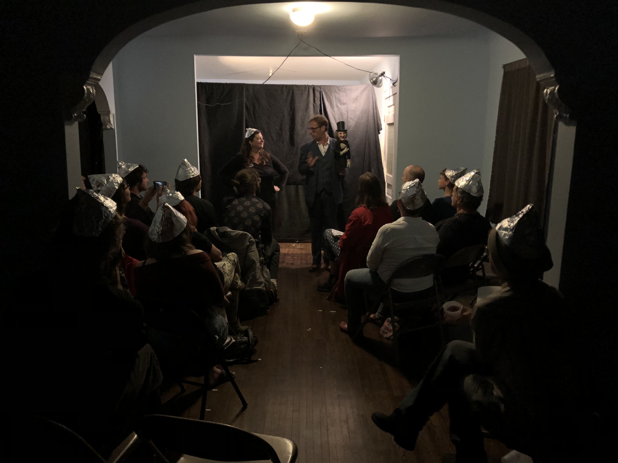 An audience seated and wearing handmade aluminum foil hats, while watching an indoor performance that includes live actors and a hand puppet with a top hat.