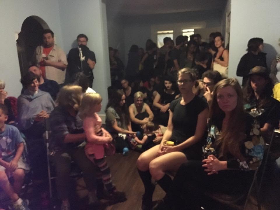 A large indoor audience of adults and children, densely packed with some seated and some standing, in anticipation for the first performance act of the evening.