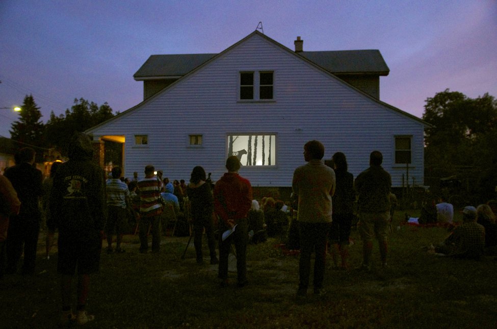 An evening outdoor shadow puppet performance, cast from the window of the Detroit Puppet Company studio, with a standing audience watching a shadow puppet performance of a tiger walking through a landscape of tree trunks.