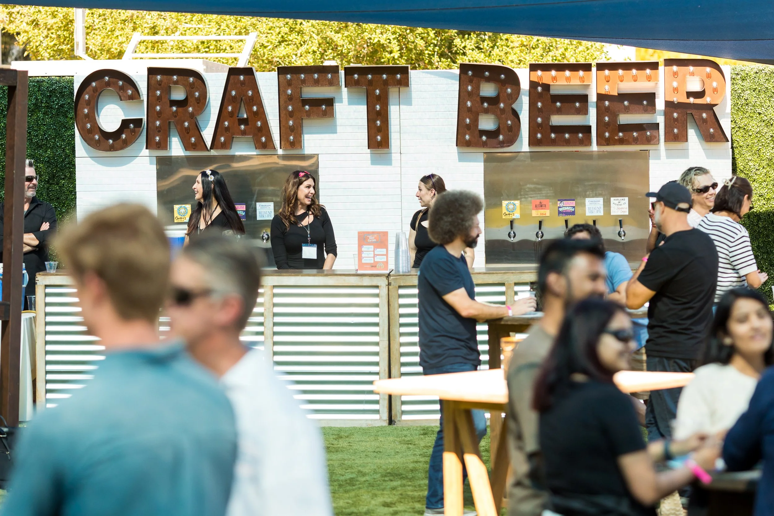 People gathered outdoors at a craft beer festival, standing near a white booth with a large lighted sign spelling 'CRAFT BEER'. Several individuals are talking and smiling near the booth, with others in the foreground and background enjoying the event.