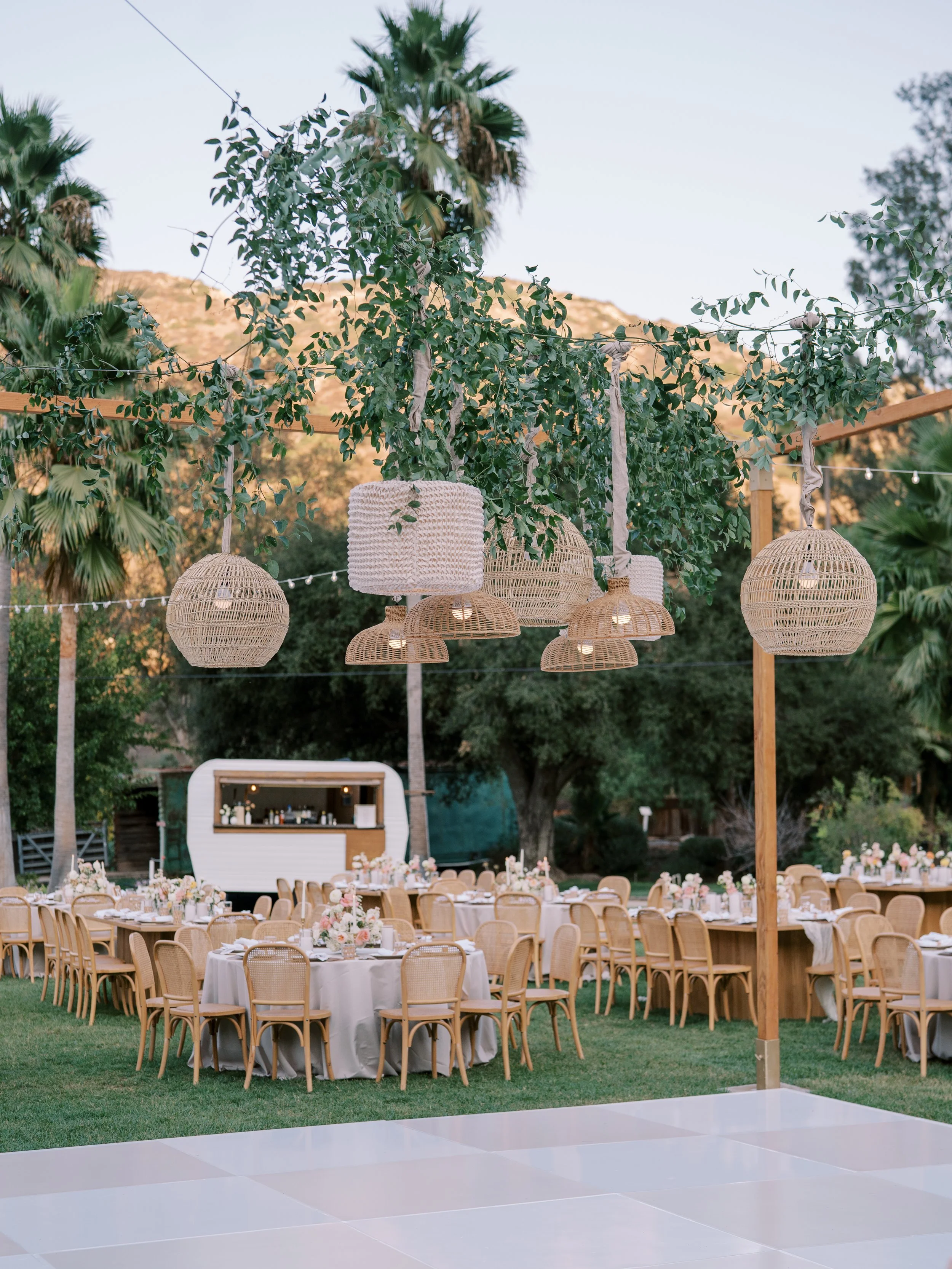 Outdoor wedding reception setup with round tables covered in white tablecloths, surrounded by wooden chairs, decorated with floral centerpieces, set on a grassy area. Hanging lanterns and string lights are suspended from wooden beams and trees, with a backdrop of tall bushes and trees, and a bar area in the distance.