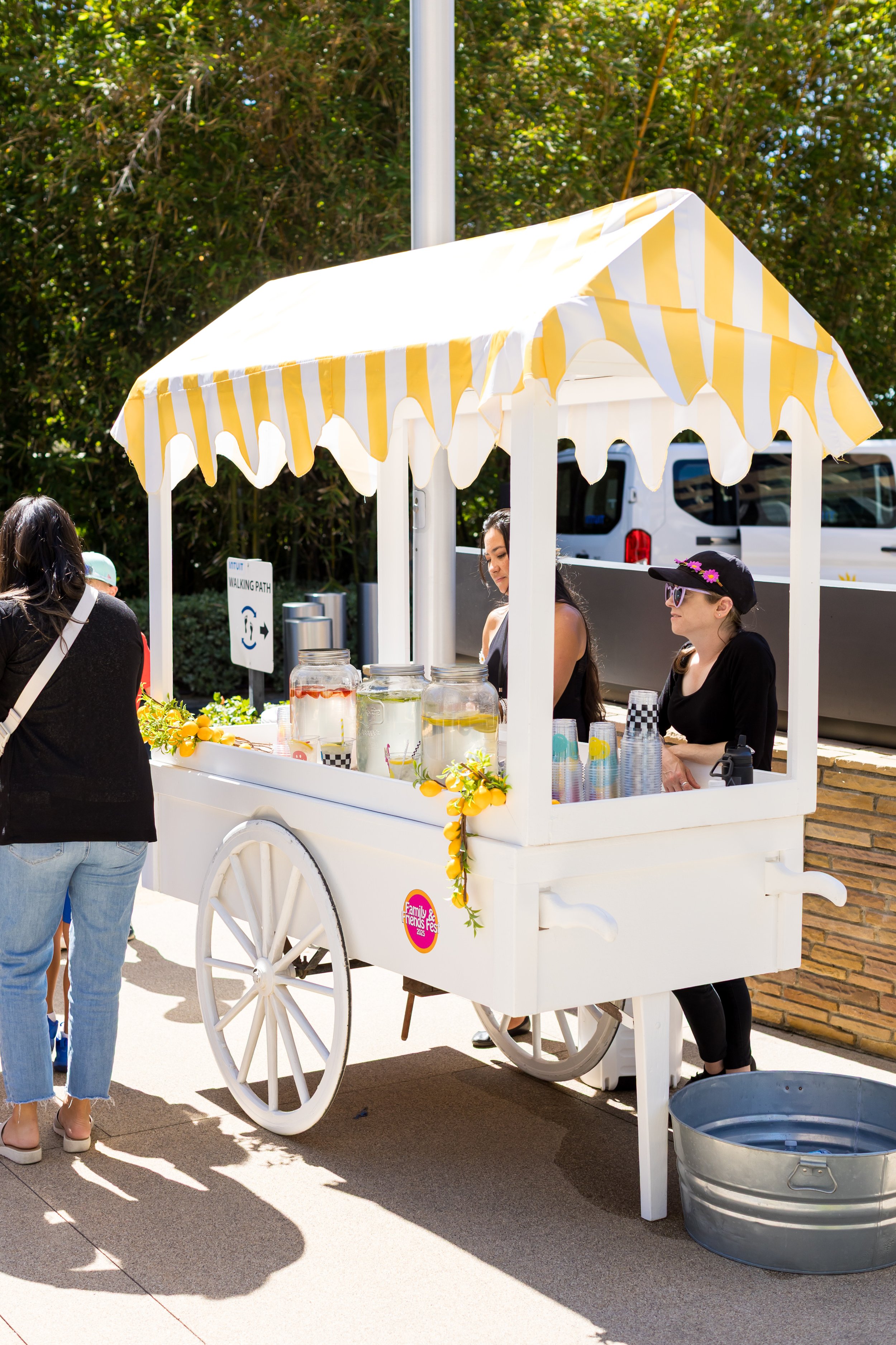 A white vintage-style ice cream cart with yellow and white striped canopy, decorated with yellow lemons and green foliage, set outdoors with people around. There are jars on the cart and a sign indicating a walking path nearby.