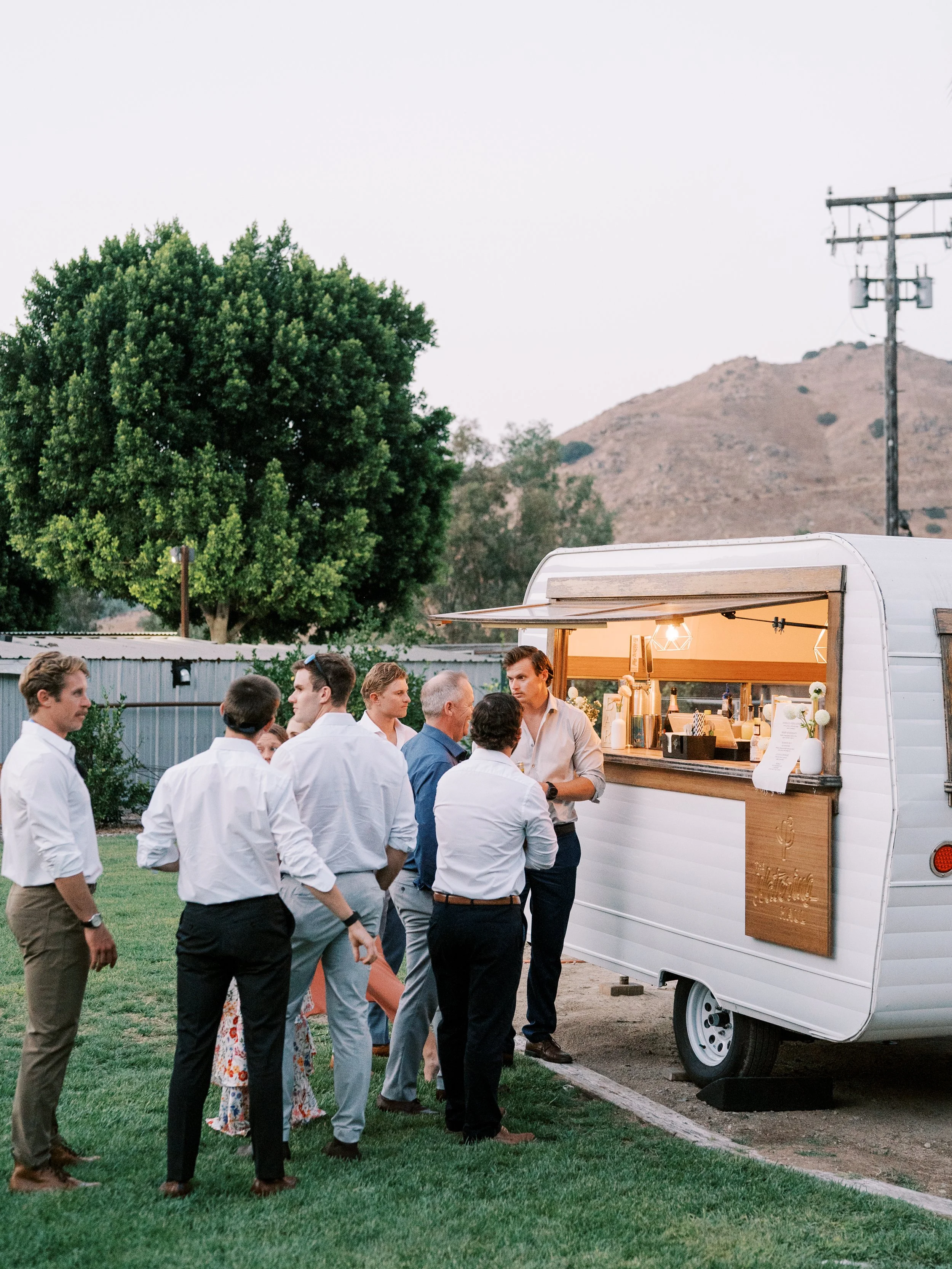 Group of people in business attire gathered outside near a mobile bar trailer, engaging in conversation during an outdoor event, with a tree, mountains, and utility pole in the background.
