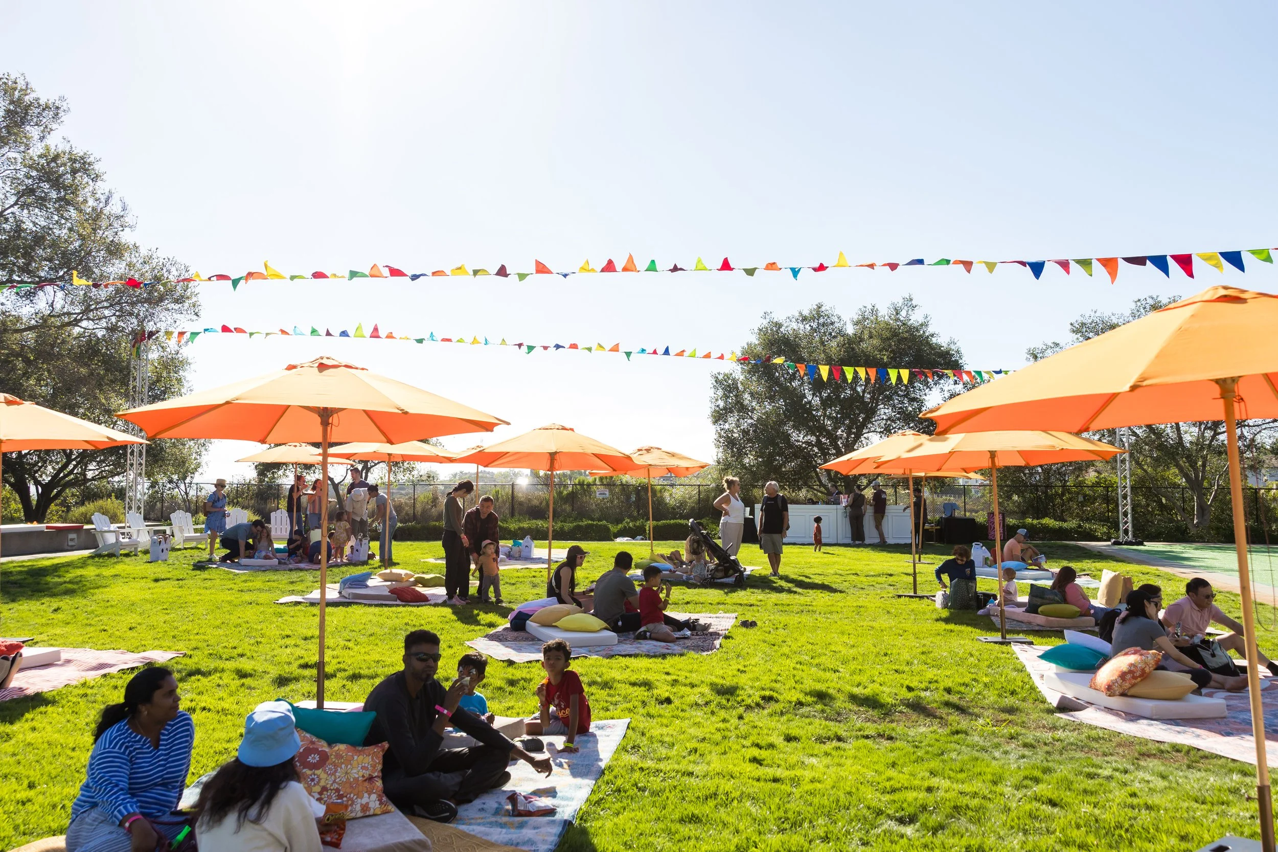 People sitting and relaxing on blankets and cushions on a grassy outdoor area during a sunny day, with orange umbrellas providing shade, colorful bunting flags overhead, and trees in the background.