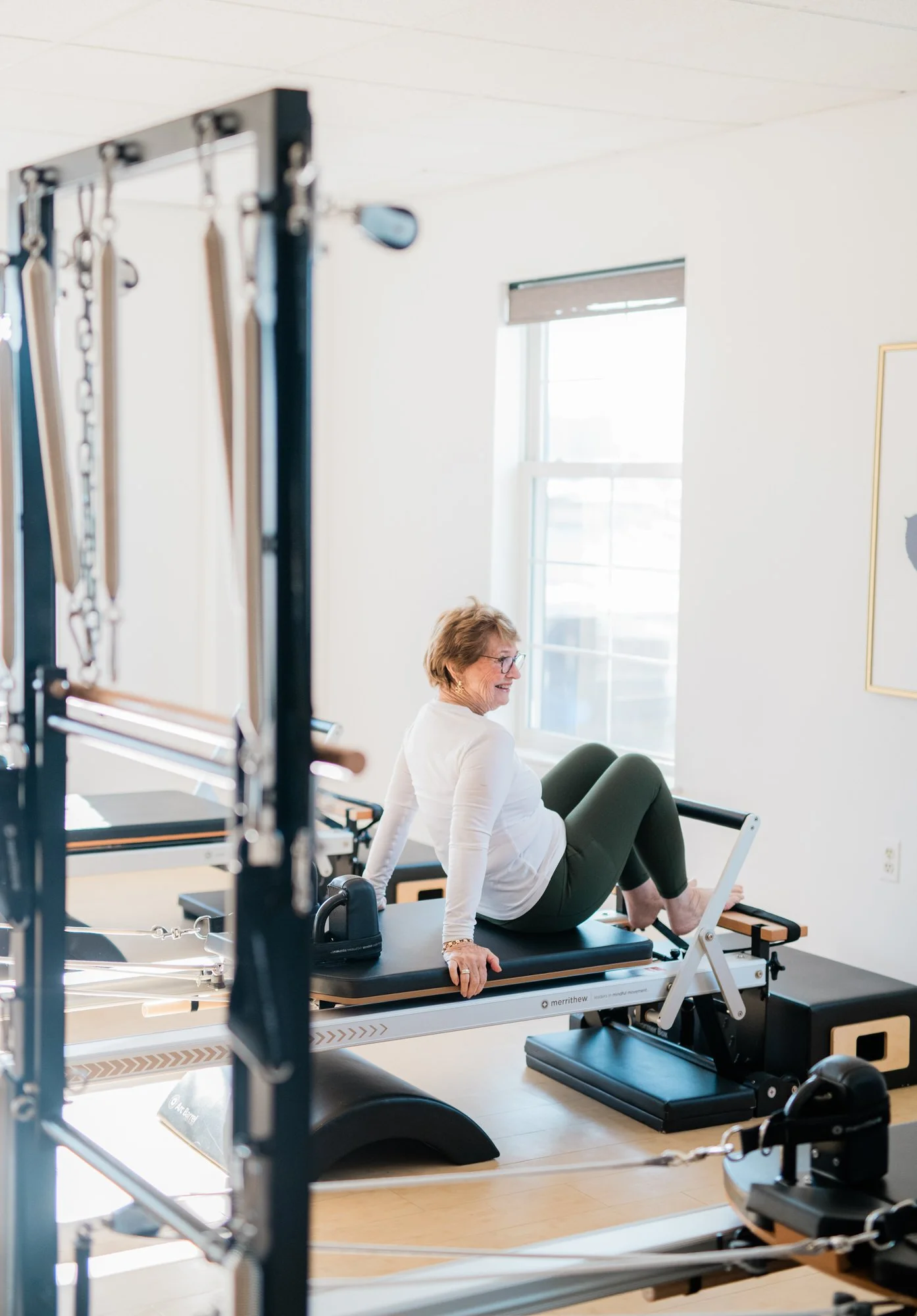 A woman with short light brown hair and glasses sitting on a Pilates reformer machine in a bright room with a large window, smiling.
