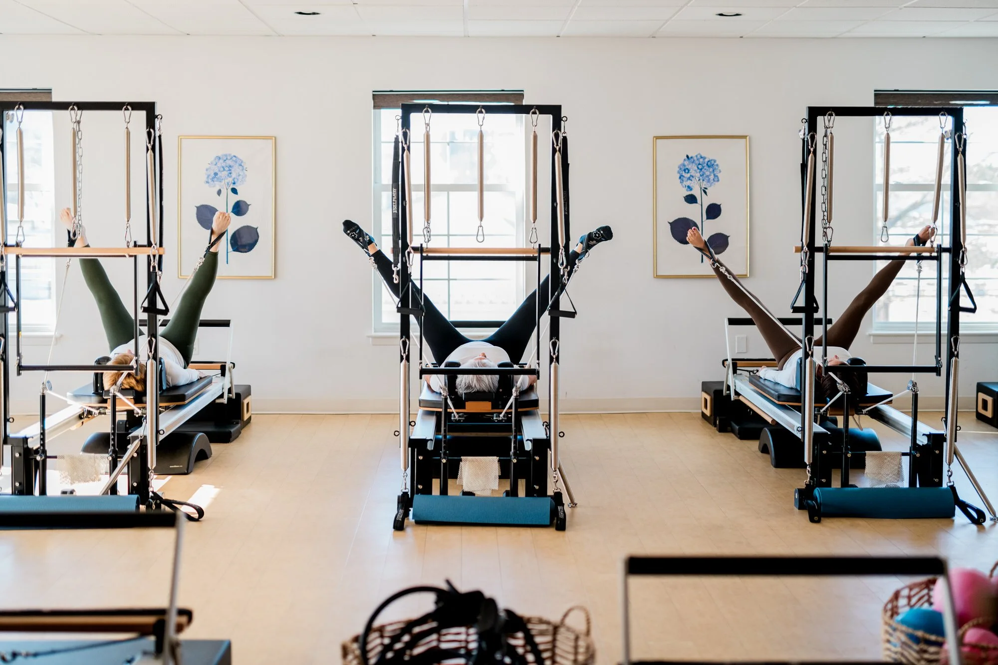 Three people lying on Pilates reformer machines at The Pilates Place in Timonium, stretching with their legs in the air. The studio has large windows, framed floral artwork on the walls, and light-colored hardwood floors.